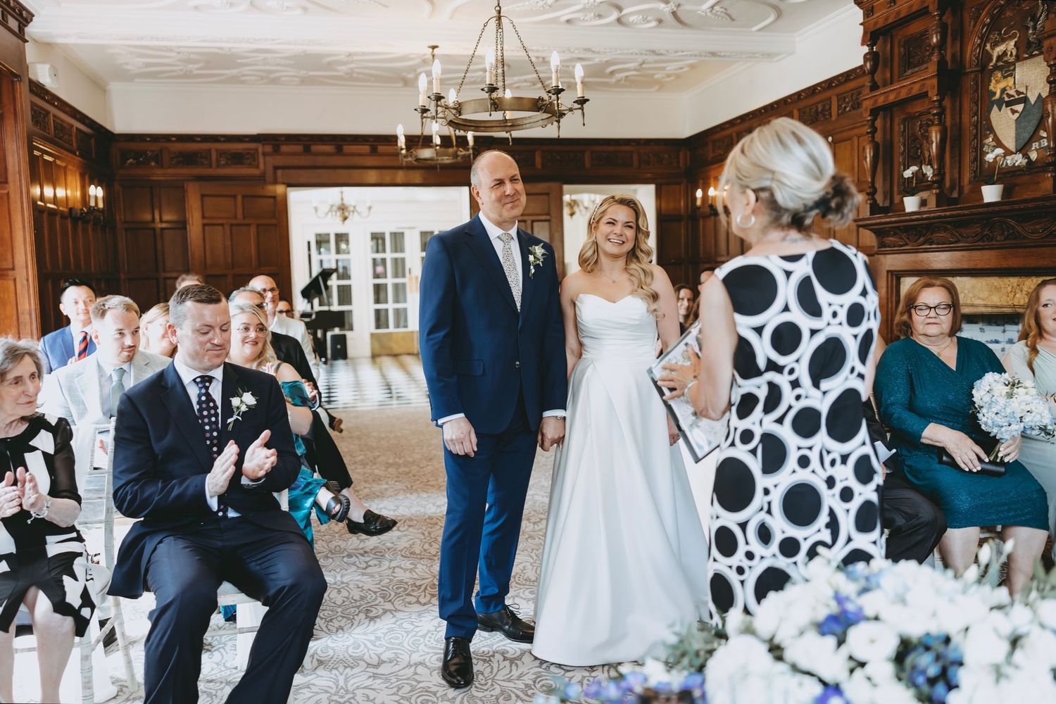 A wedding ceremony with guests watching in a traditional wood-paneled room with chandelier.