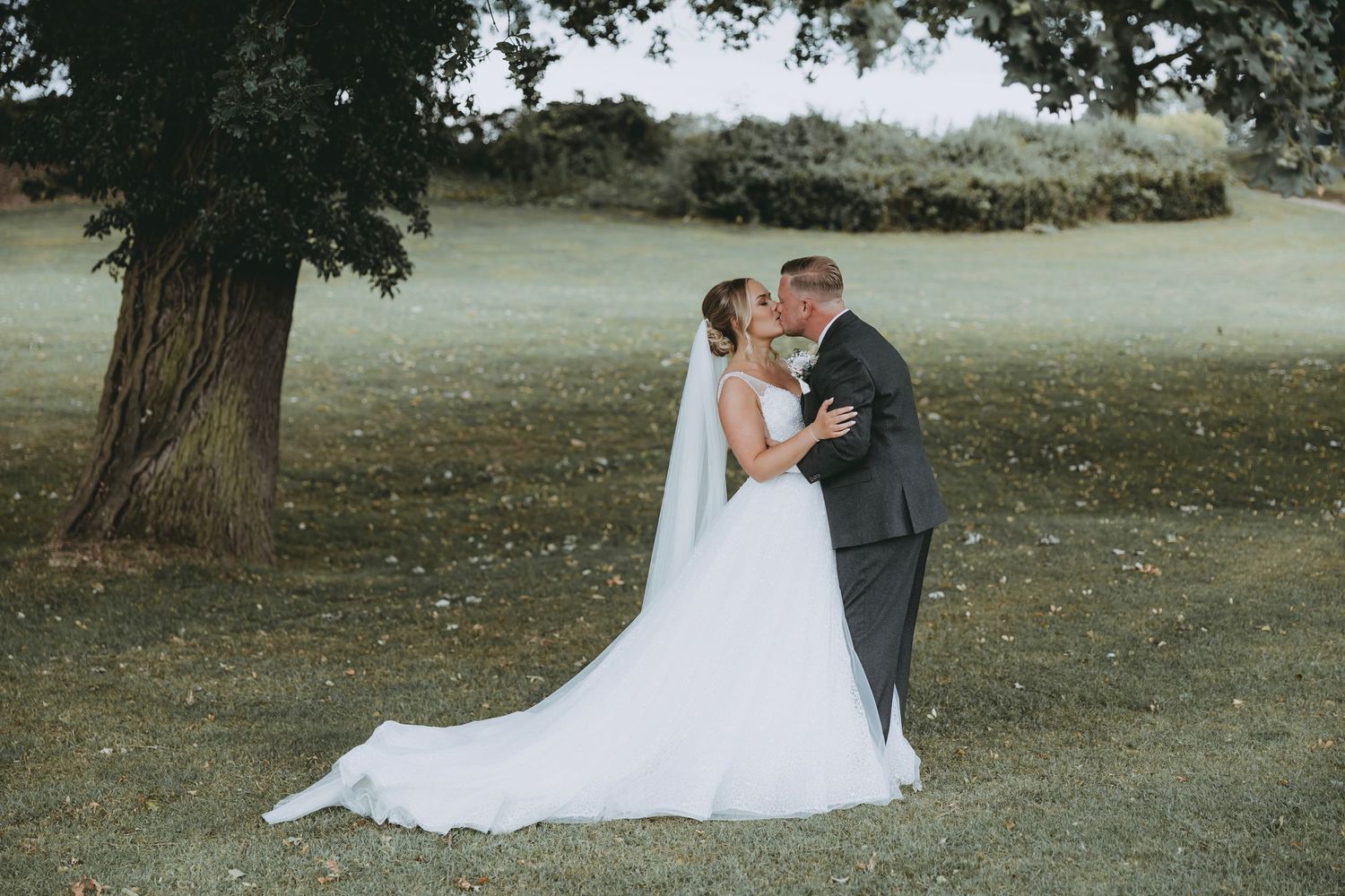 Newlywed couple embraces on a grassy lawn with trees in the background.