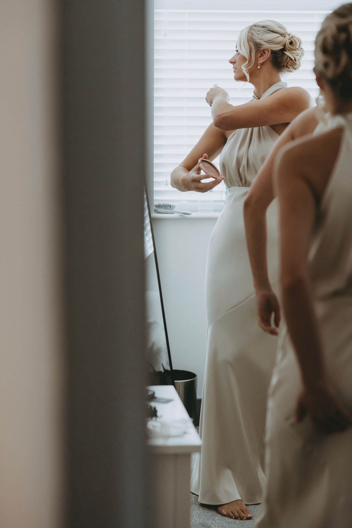 Wedding preparations continue as attendants in white dresses gather in a sunlit room.