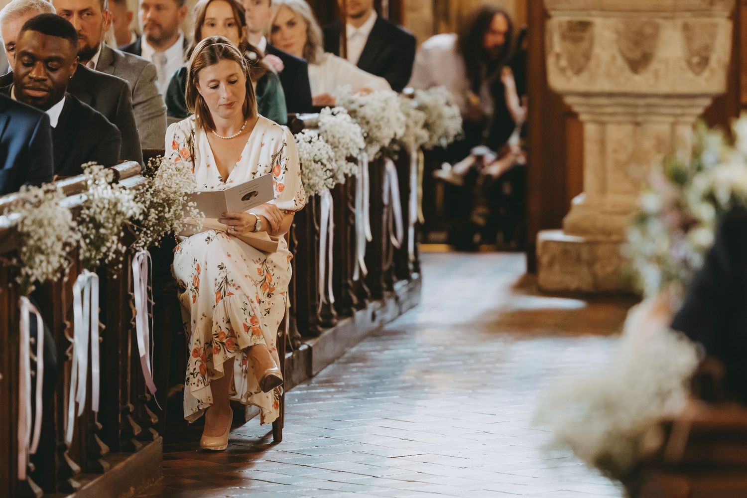 Wedding guests line both sides of a church aisle decorated with white floral arrangements and candles.