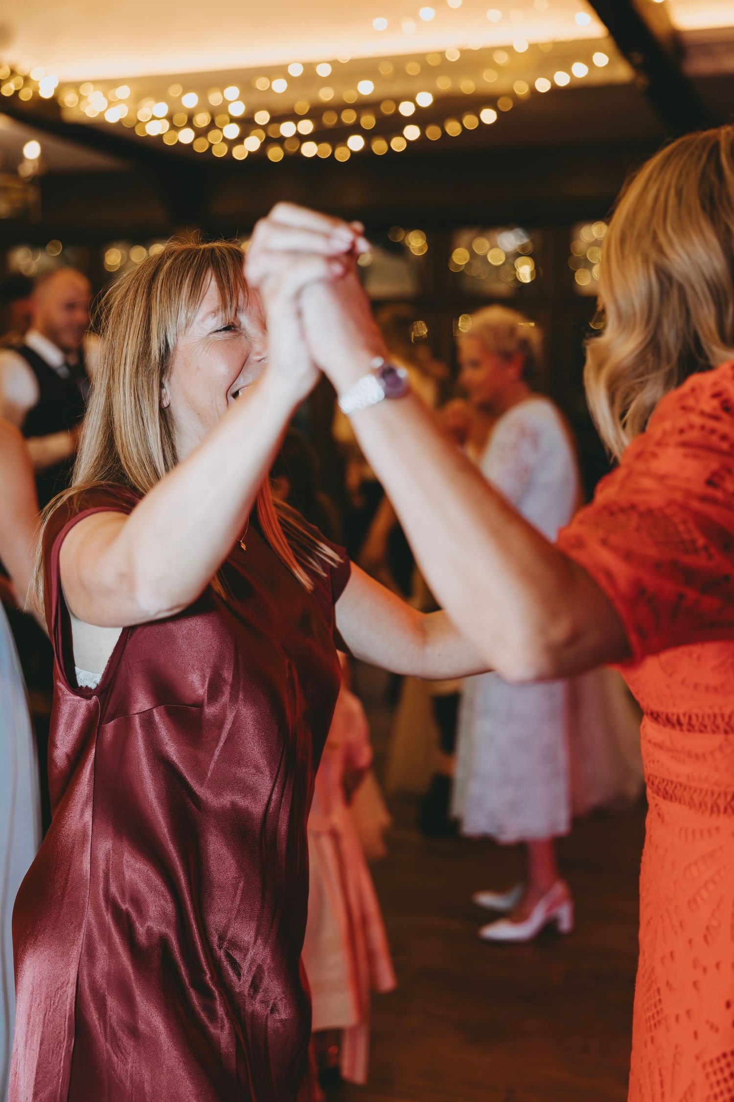 Wedding party members dance in coral and burgundy dresses under string lights.