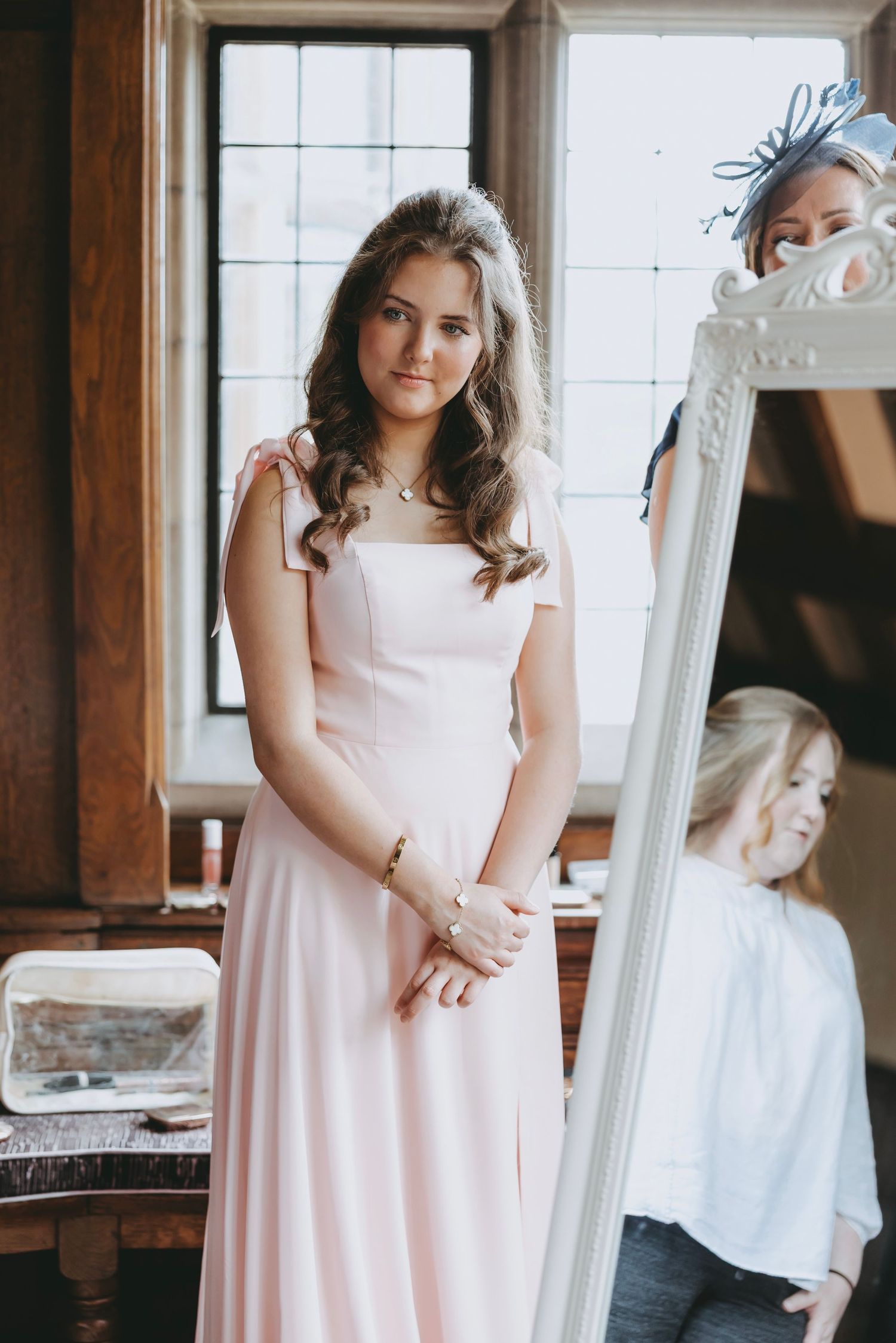 Bridesmaids in pink dresses pose near a vintage white mirror in natural light.