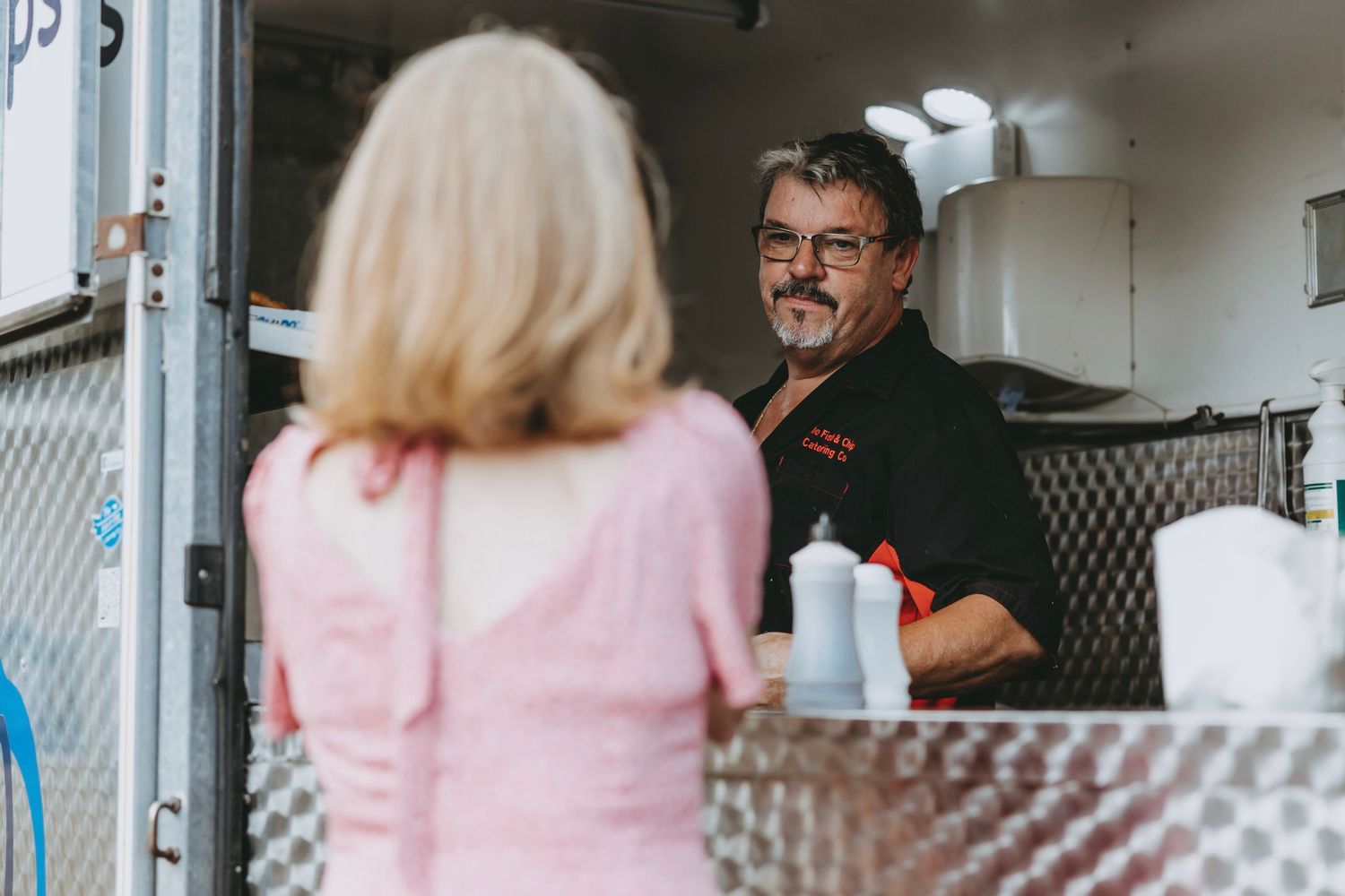 A person in a pink shirt stands in a commercial kitchen while someone works in the background.