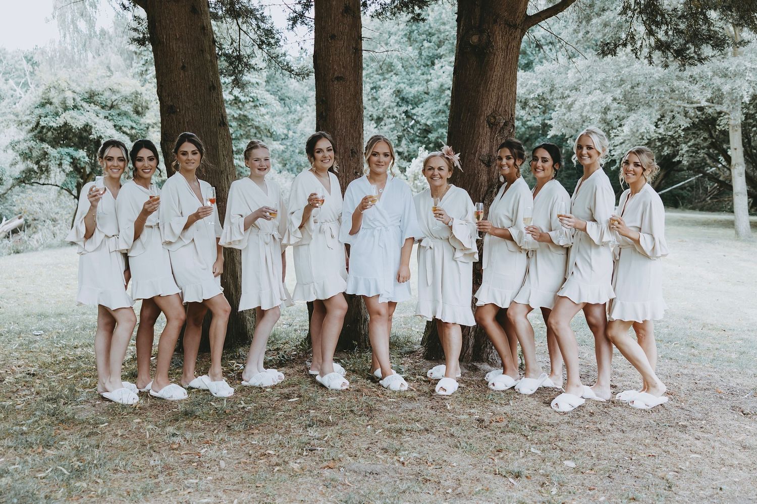 Bridal party in matching white robes posing together outdoors under trees.
