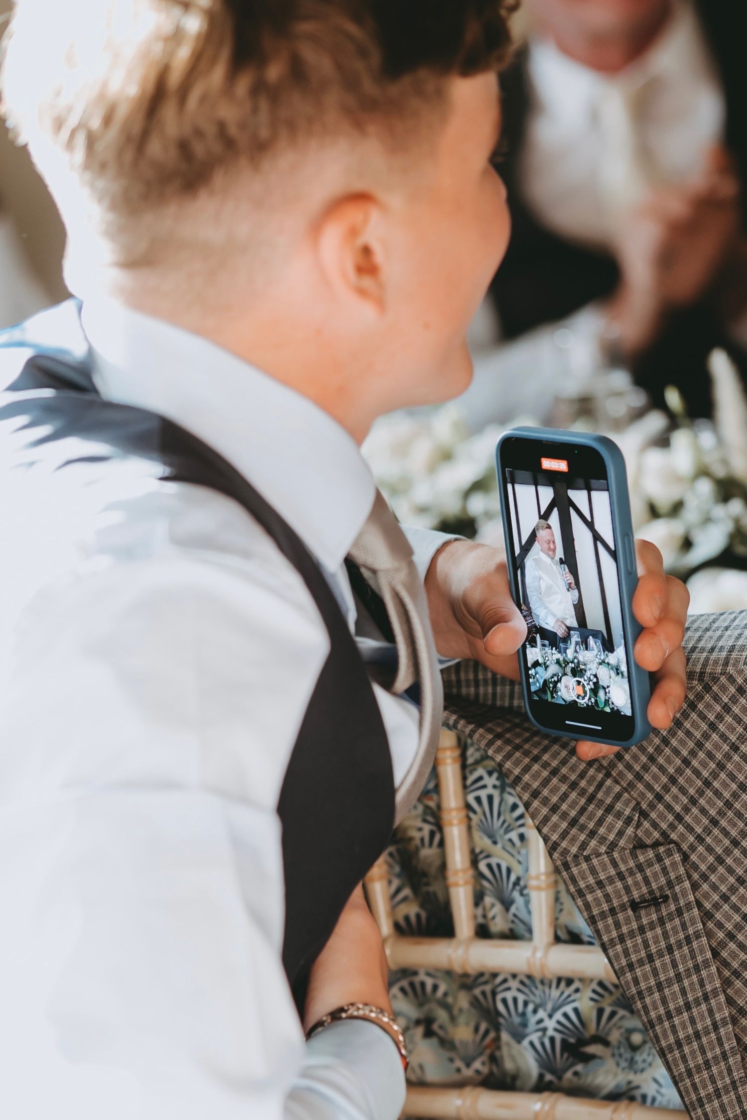 Young guest in formal attire uses smartphone to capture wedding reception moments.