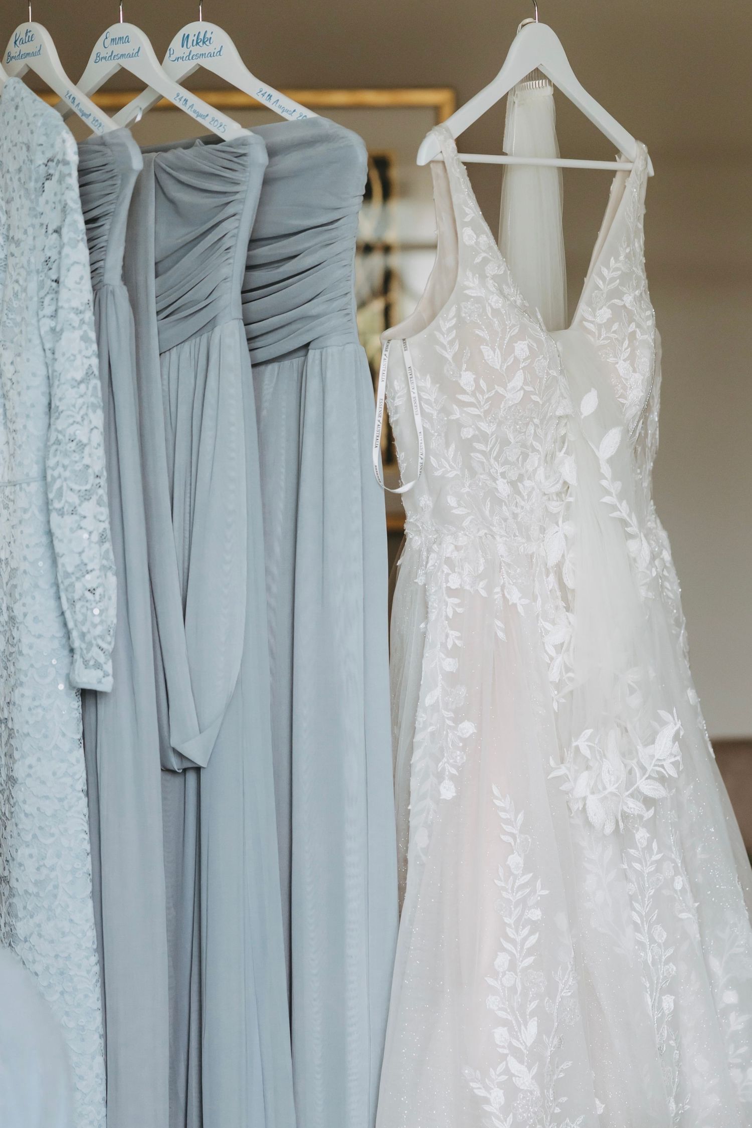 Wedding dresses and light blue bridesmaid gowns hanging on white hangers against a neutral wall.