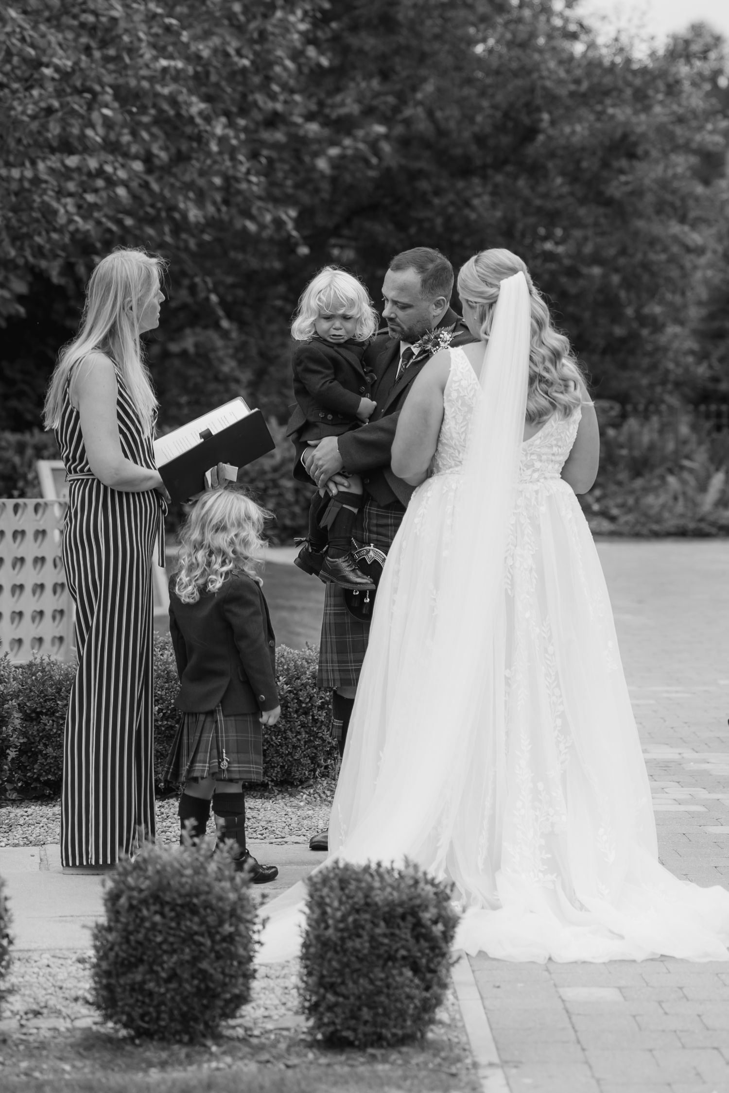 Black and white wedding photo showing guests in Scottish formal wear on stone patio.