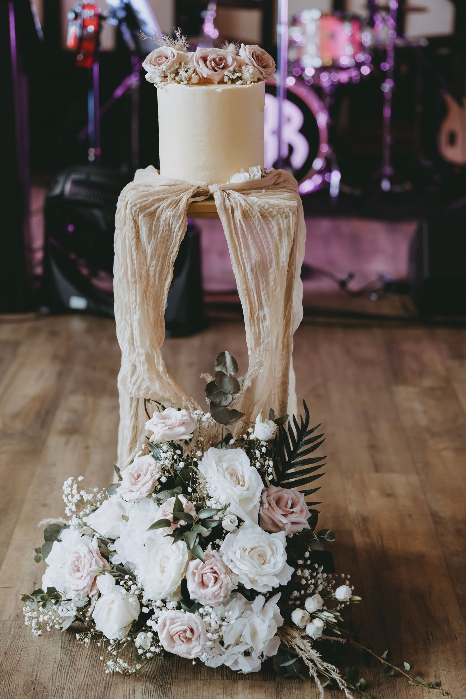 Elegant white wedding cake with draped fabric and pink rose floral arrangement on wooden table.