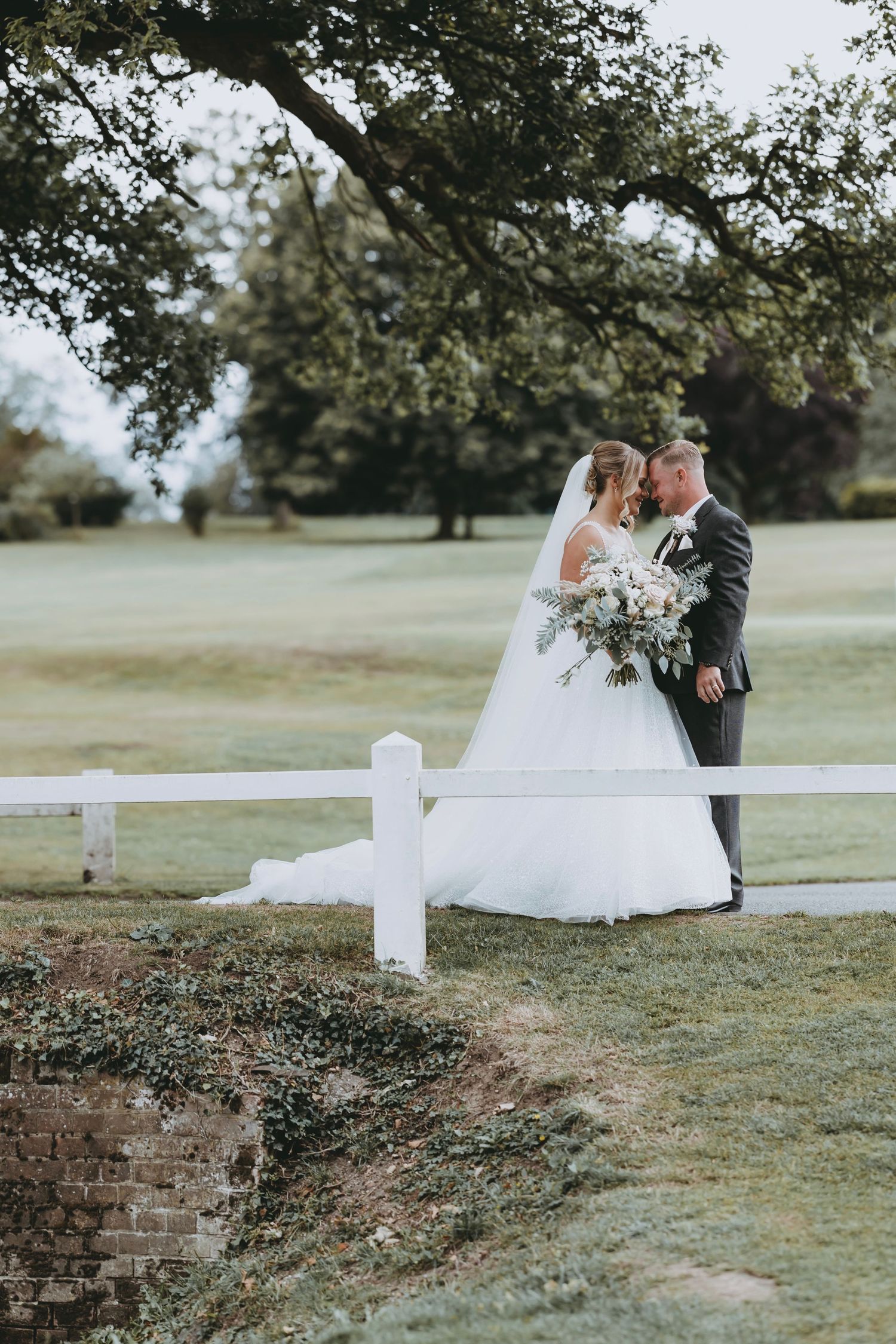 Elegant wedding couple poses together on grassy hillside with white fence and trees in background.