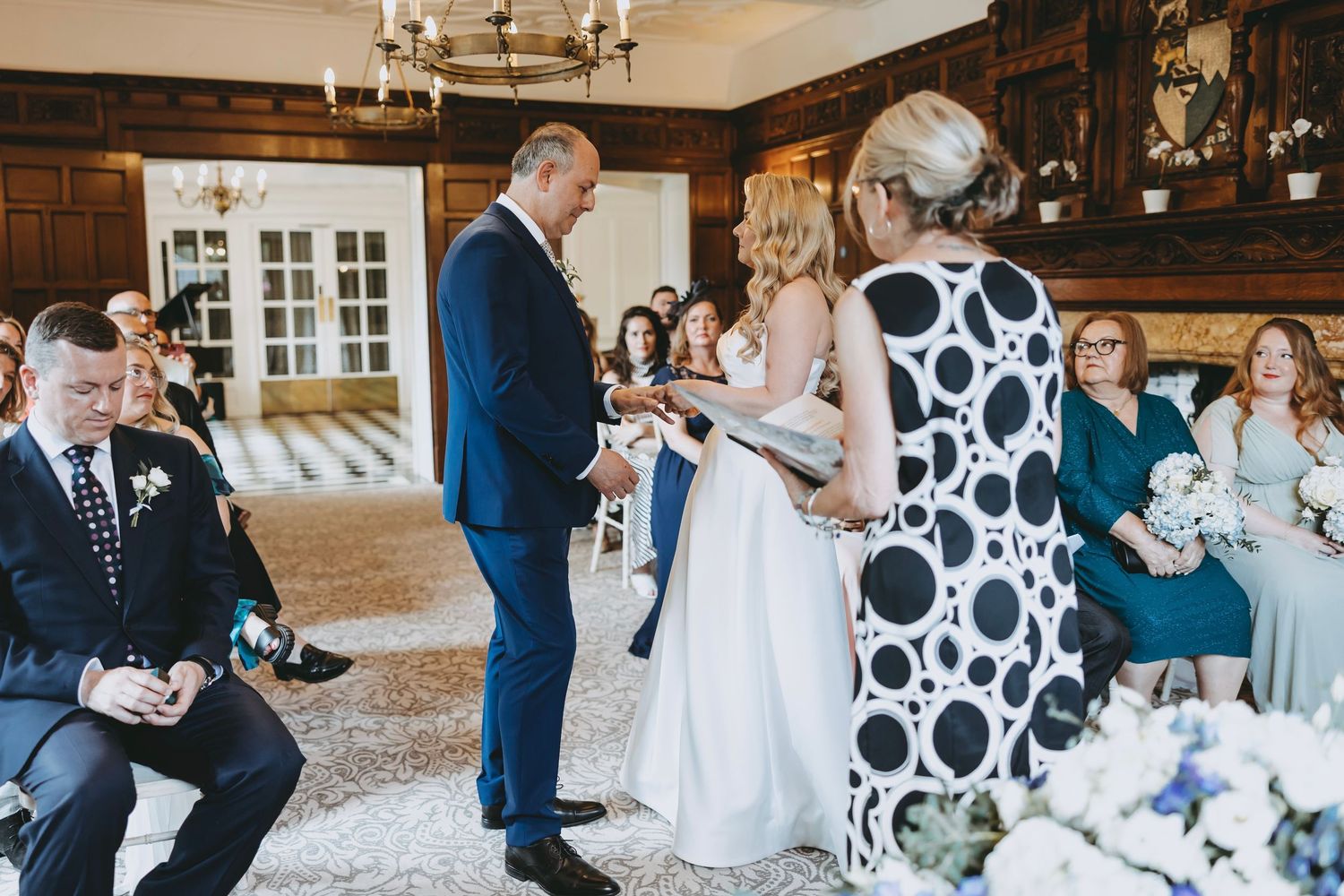 Wedding ceremony participants stand in a wood-paneled room with chandeliers and polka dot decorations.