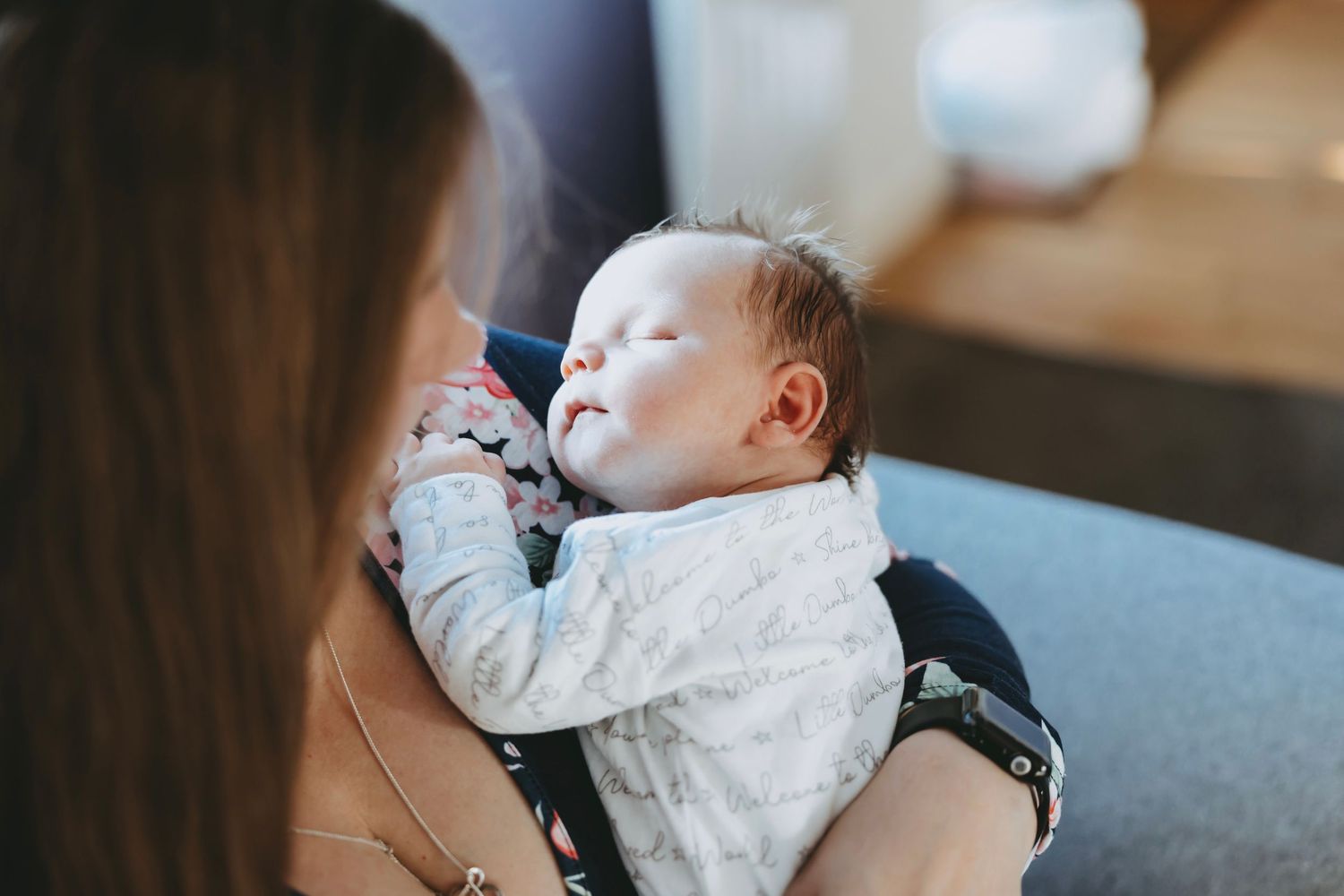 Baby in white lace dress yawning while being held in someone's arms on a sofa.