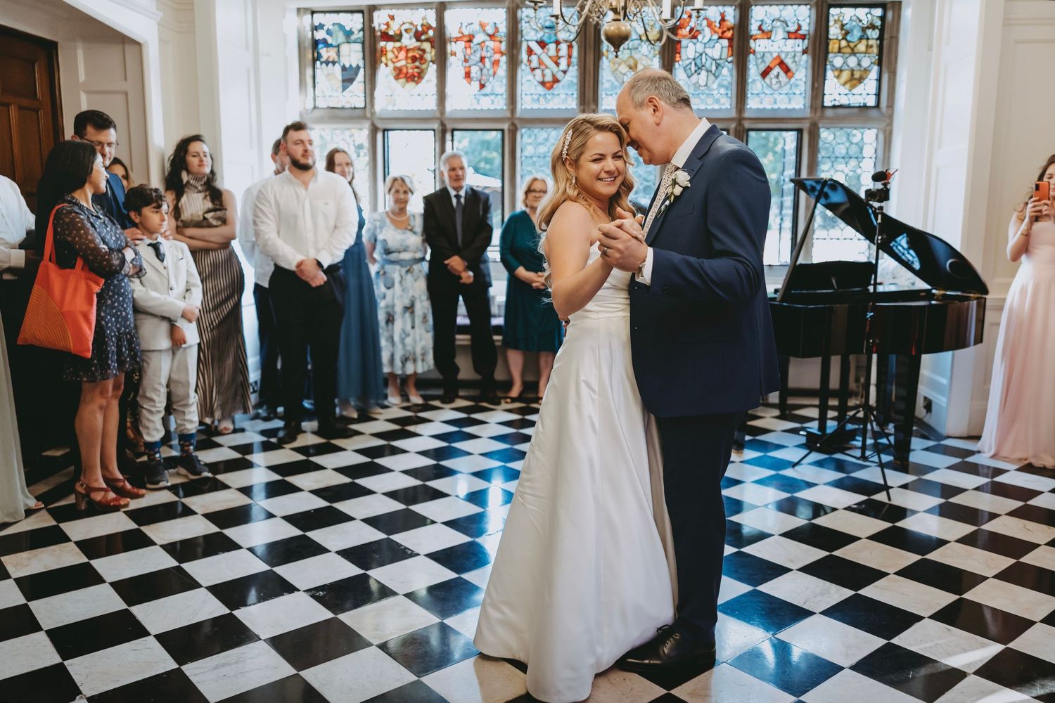 Wedding celebration with dancing couple on black and white tile floor beneath ornate windows.