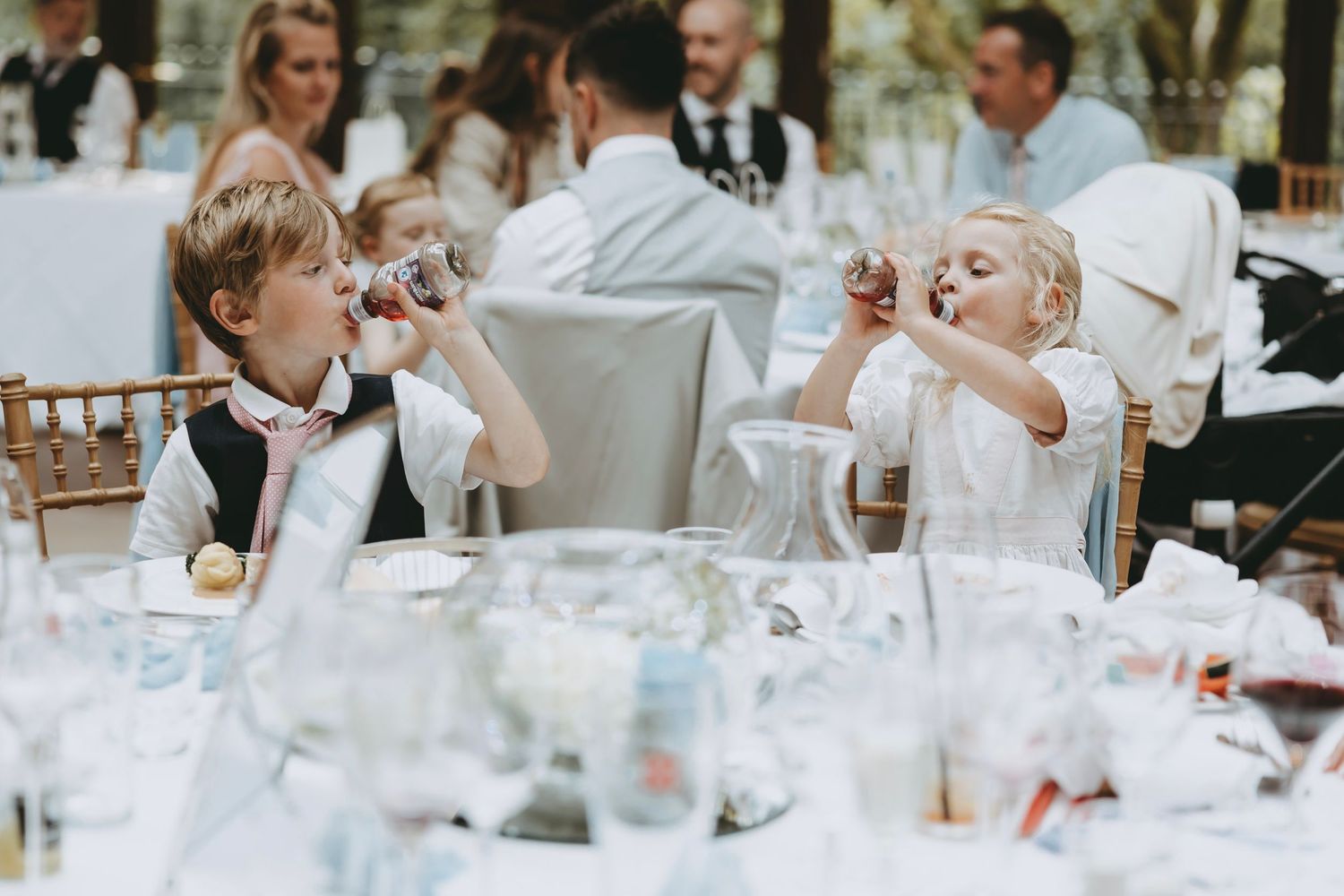 Two children in formal attire drink from glasses at an elegant reception table.