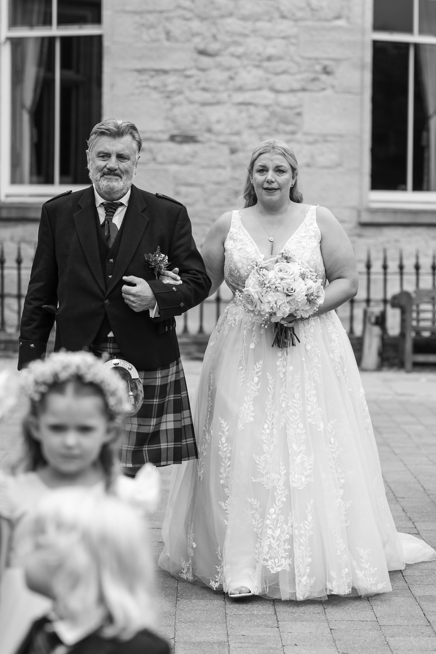 Wedding party members process down aisle during outdoor ceremony at stone building.