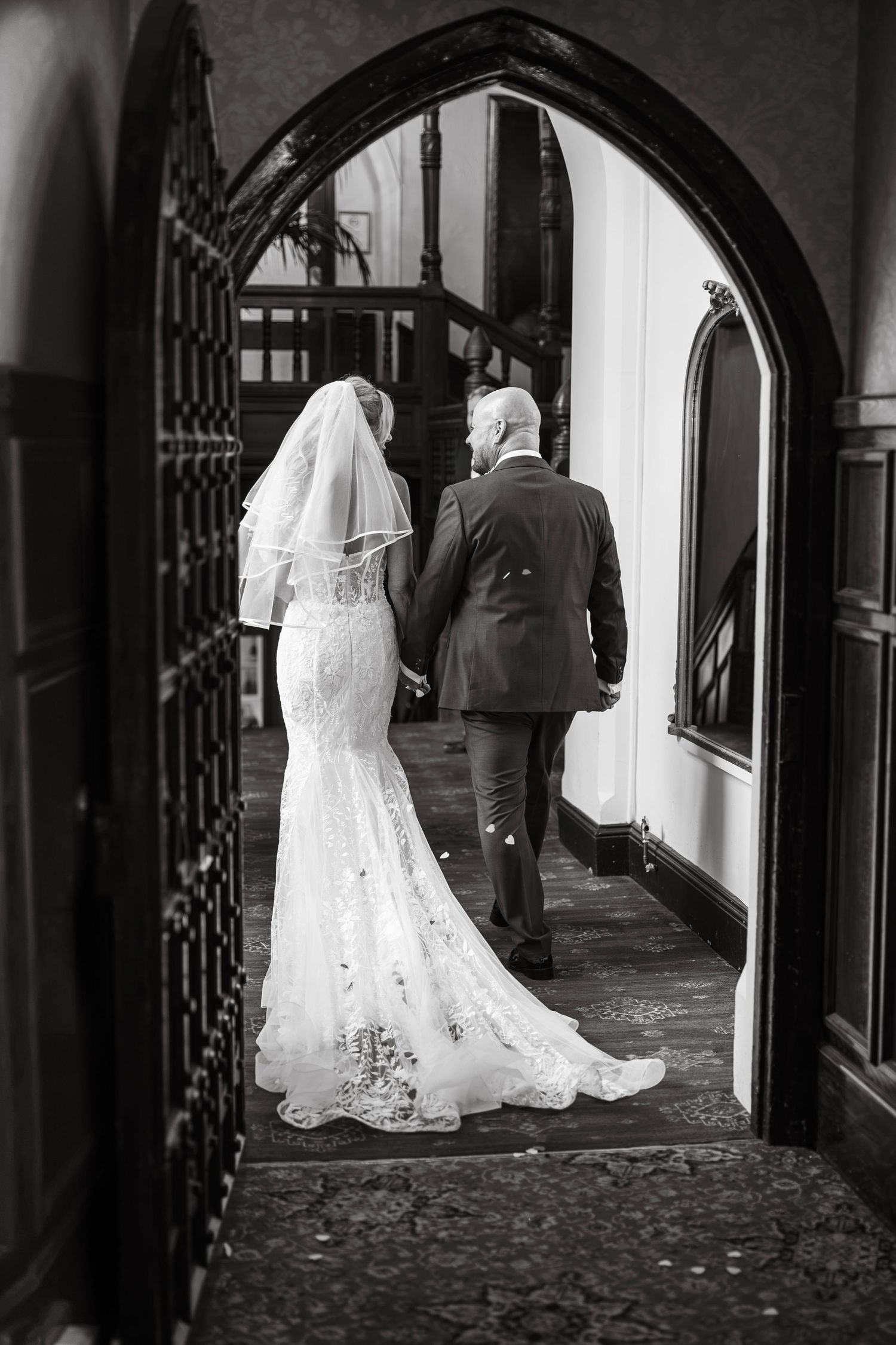 Black and white photo of a bride in lace gown and veil walking through gothic arch with escort.