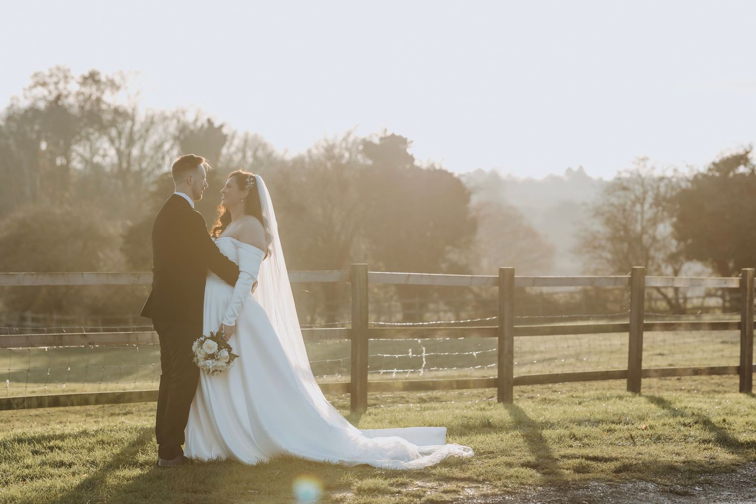 Silhouettes of a wedding couple against a sunlit countryside with wooden fencing.