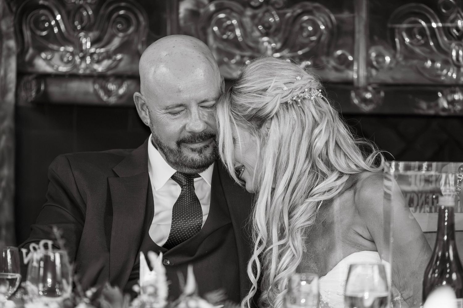 Black and white photo of a bald groom in a suit sharing an intimate moment with a blonde bride at the reception table.