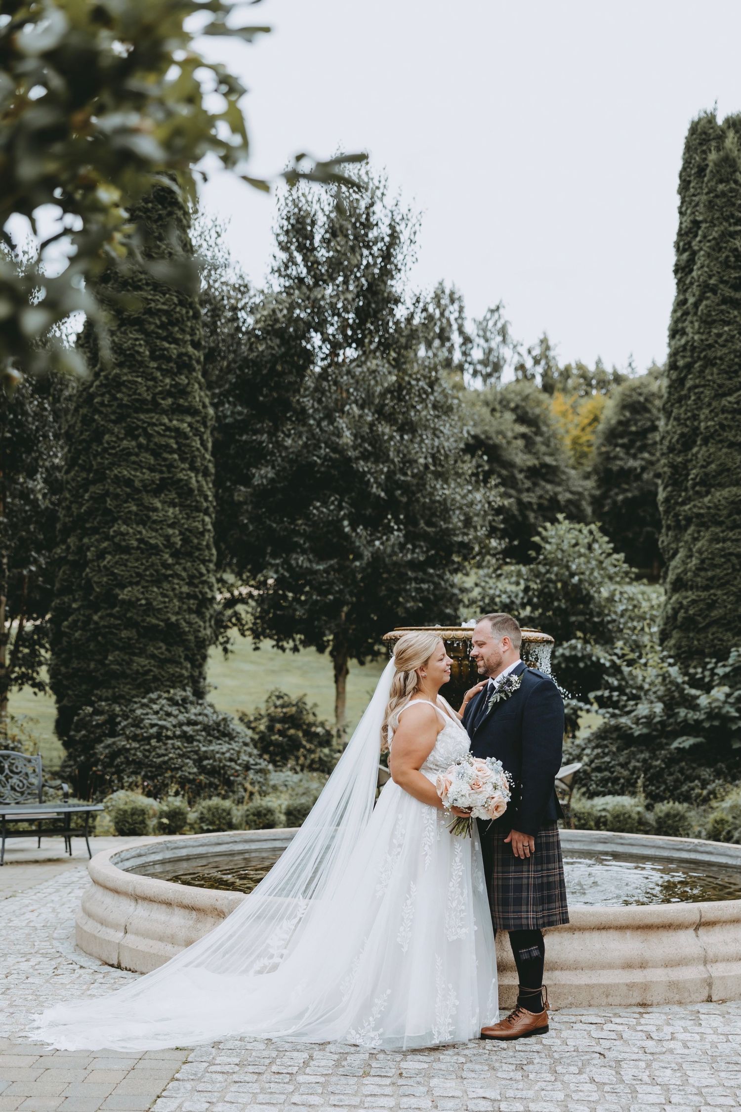 Garden wedding scene with couple in white dress and Scottish kilt near ornate fountain.