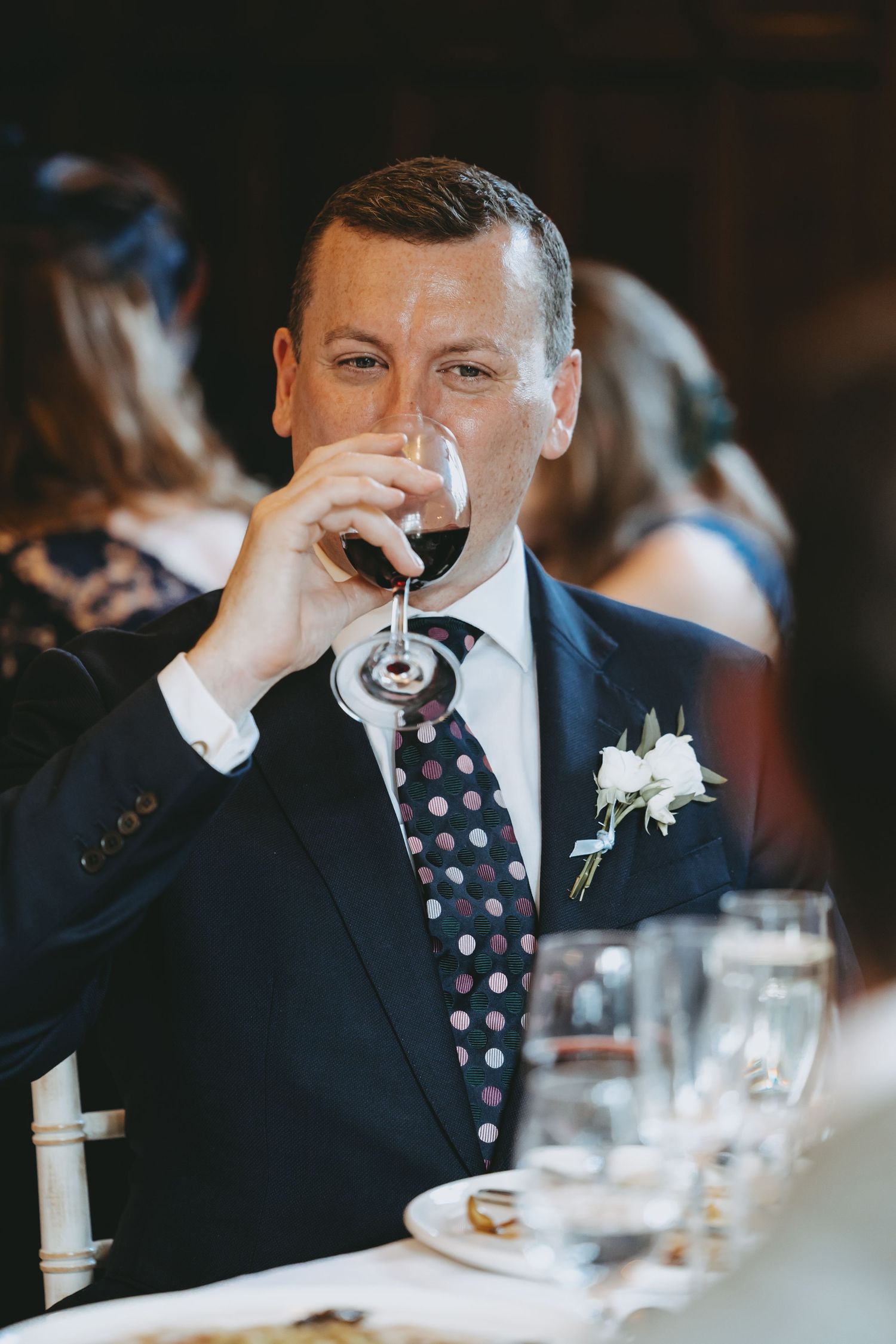 Wedding guest in dark suit and polka dot tie taking a sip from wine glass.