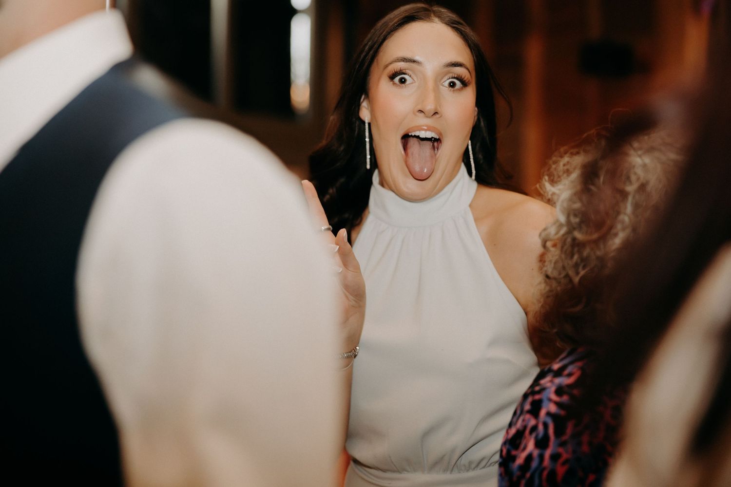 A woman in a white halter dress reacts with excitement and surprise at what appears to be a wedding reception.