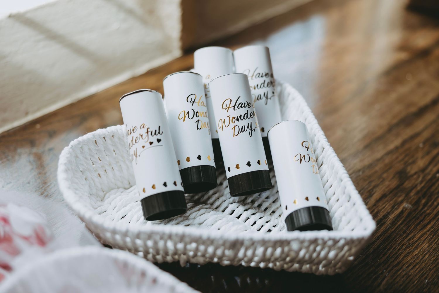 White wicker basket holding small cylindrical lip balm tubes with elegant black and gold script labels.