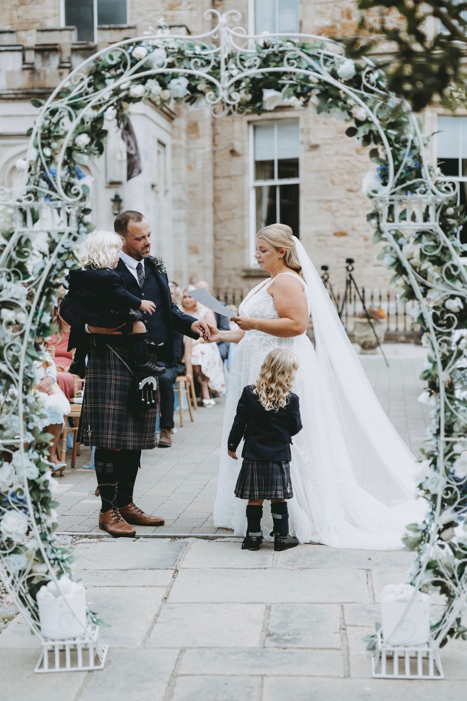 Wedding guests in traditional Scottish attire gather under a decorative floral arch during an outdoor ceremony.