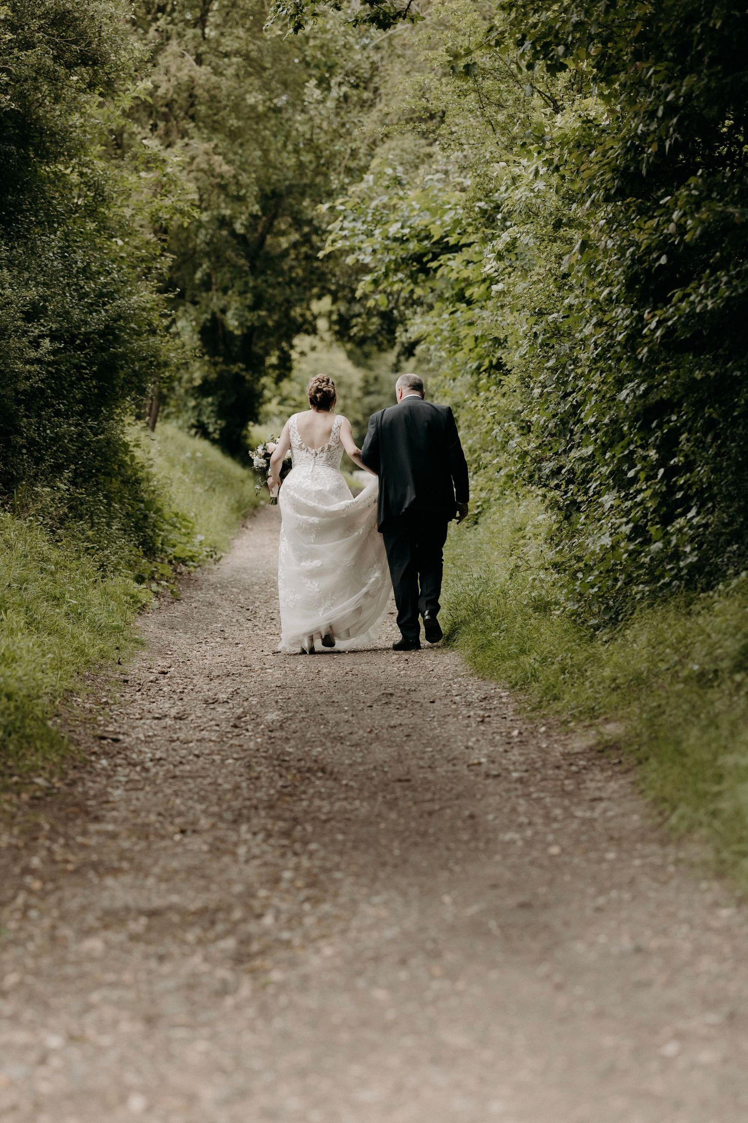 A couple walks down a tree-lined dirt path in wedding attire, photographed from behind in a romantic woodland setting.