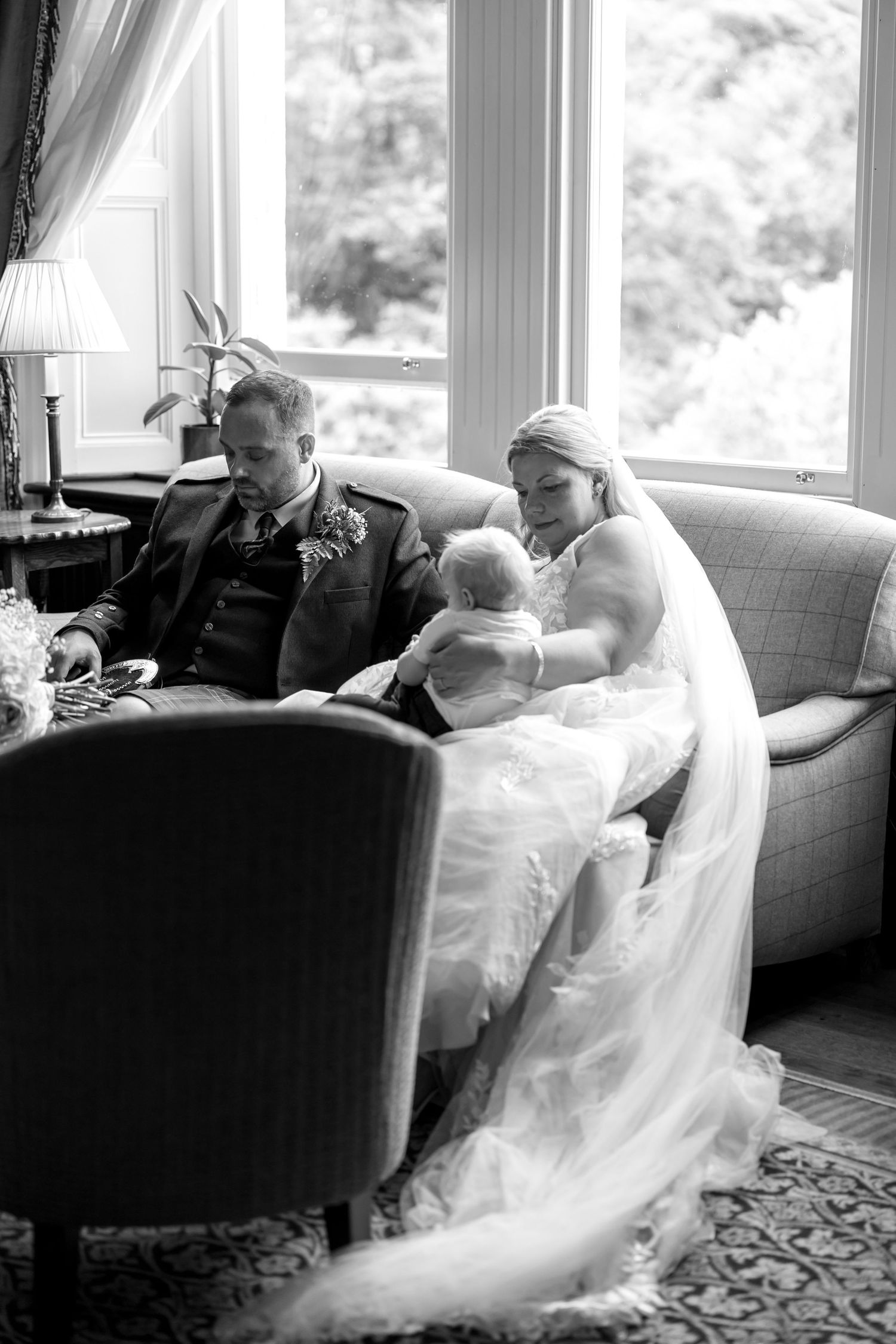 Black and white photo of wedding guests relaxing on elegant furniture by large windows.