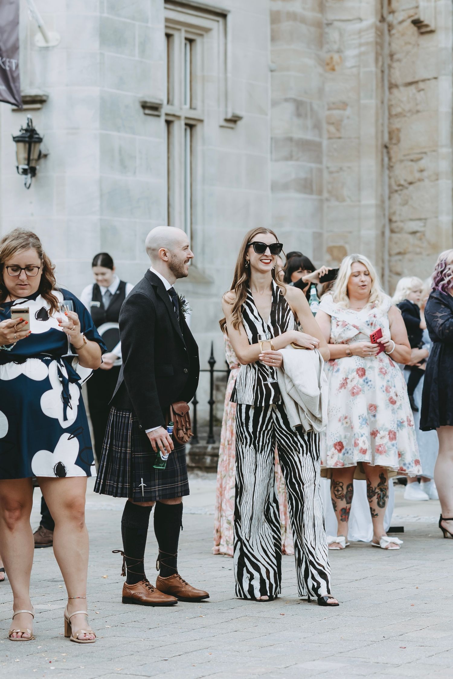 Wedding guests in stylish attire gather outside a stone building.
