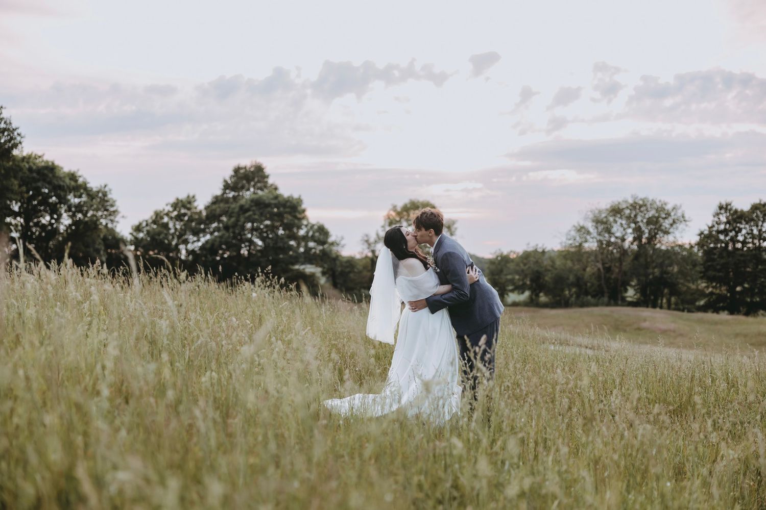 Bride and groom share a romantic embrace in a lush green meadow at sunset, with trees and pastel sky behind them.