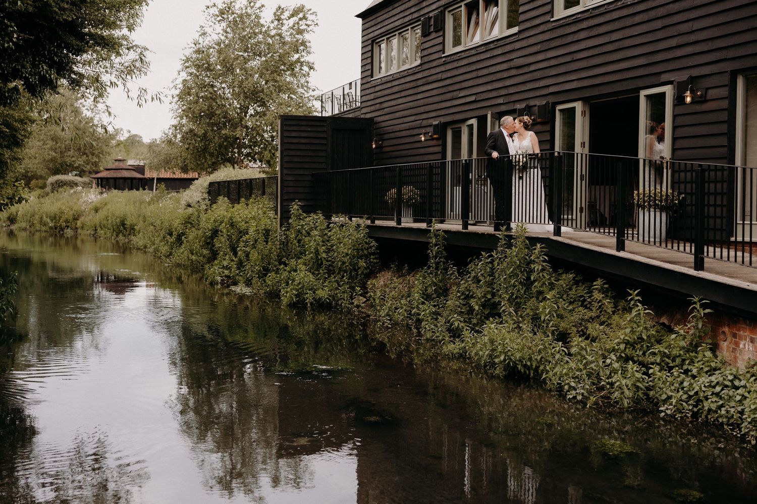 Rustic black wooden buildings line a tranquil waterway on a peaceful day at Tewinbury Farm, Hertfordshire.