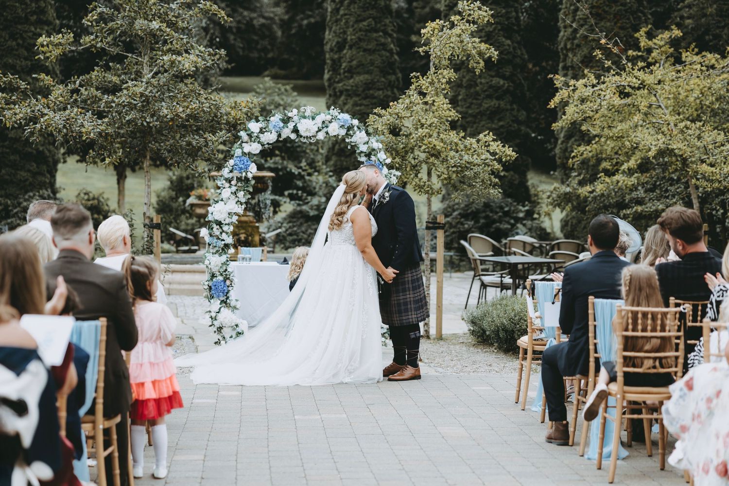 Wedding guests seated on wooden chairs at an outdoor ceremony with trees in the background.