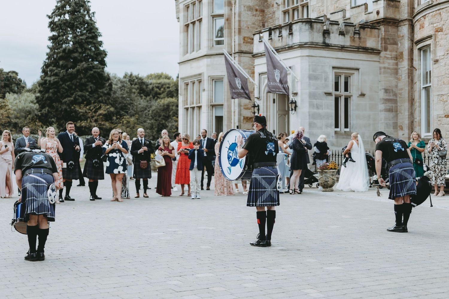 Guests and performers gather outside a grand stone building during a Scottish wedding celebration at Crossbasket Castle, Scotland.