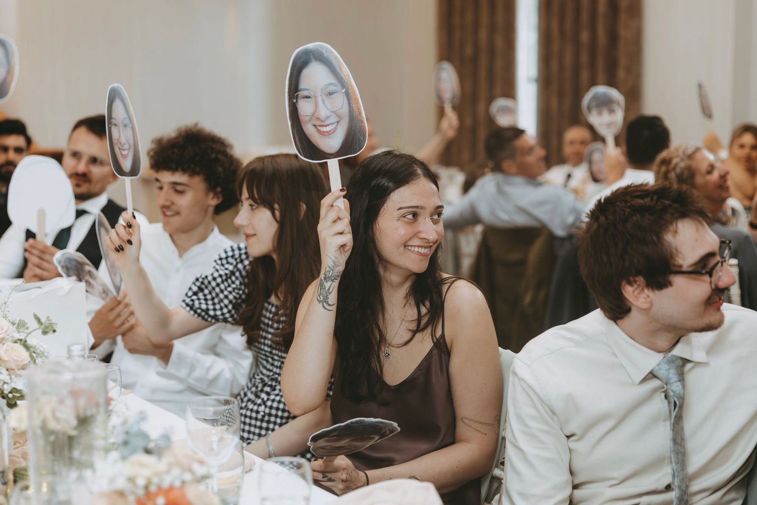 Wedding guests smiling and holding up face paddles on sticks during a fun reception celebration in an elegant venue.