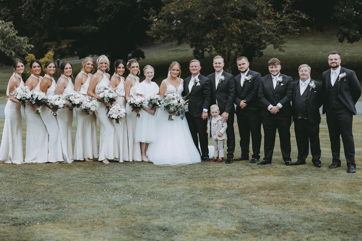 Full wedding party lineup poses outdoors with bride in white gown and attendants in formal wear.