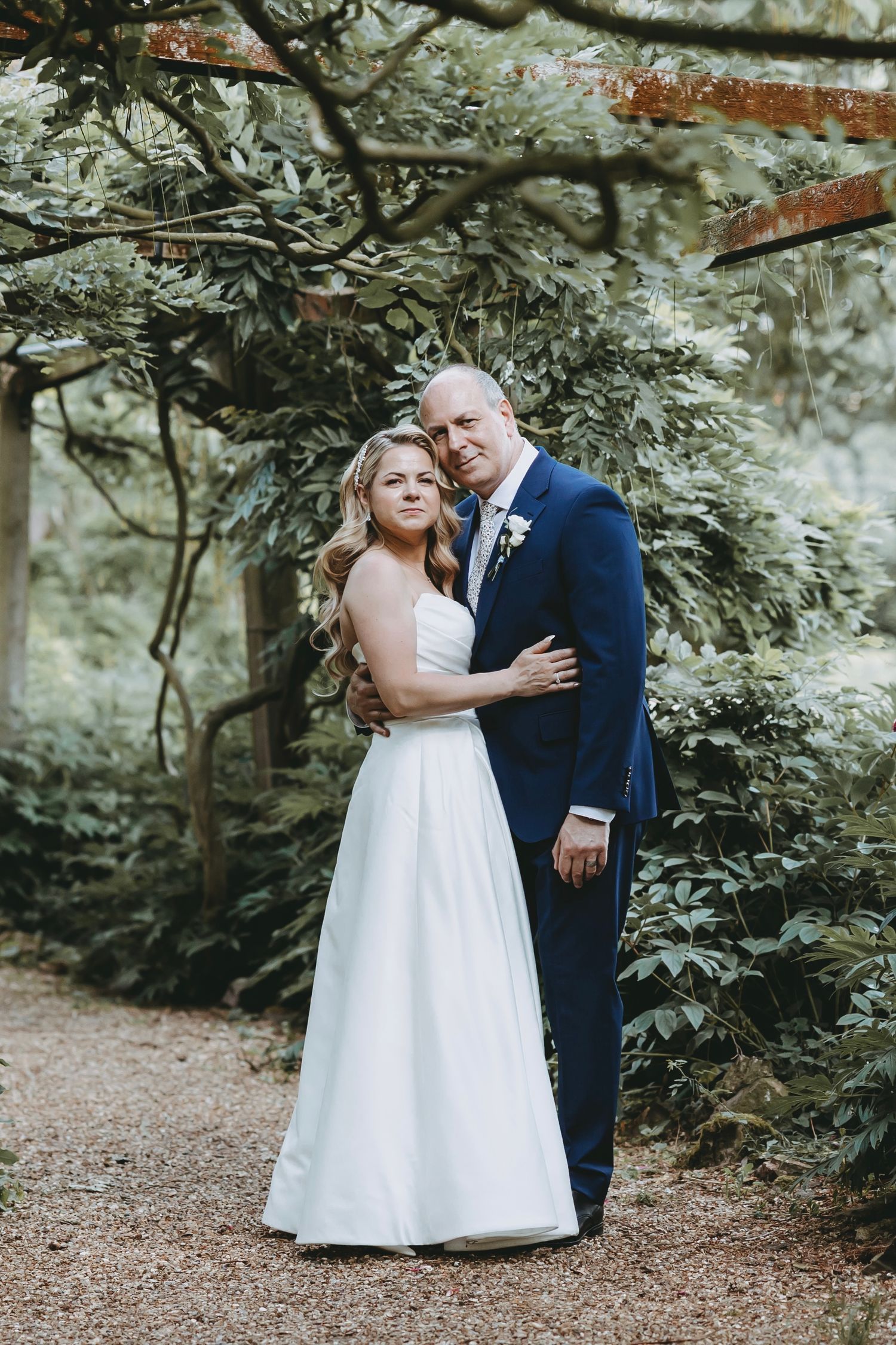 A couple in wedding attire poses together under a garden arbor surrounded by greenery.