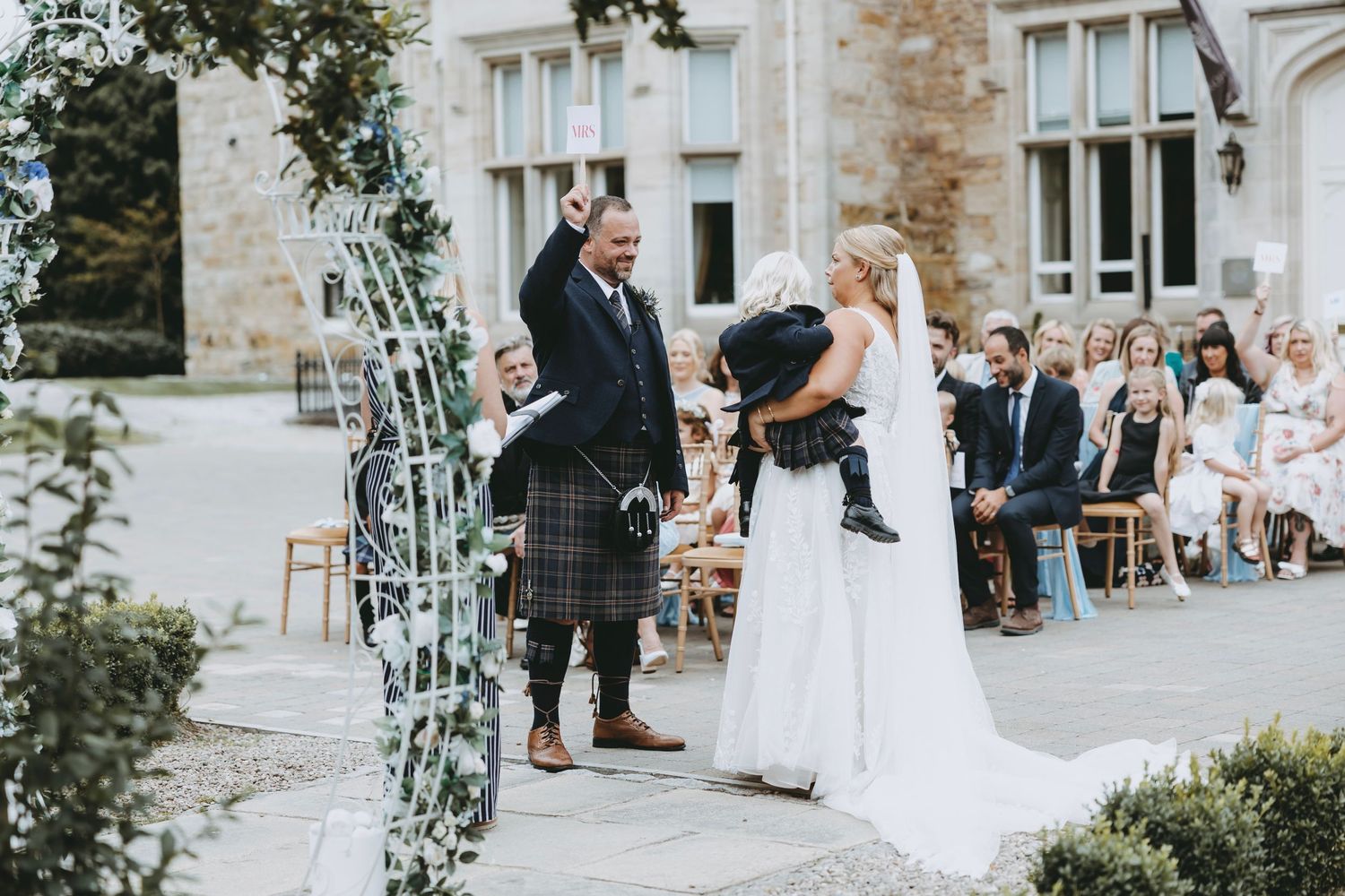 Outdoor wedding ceremony setup with floral arch and chairs against stone building backdrop.