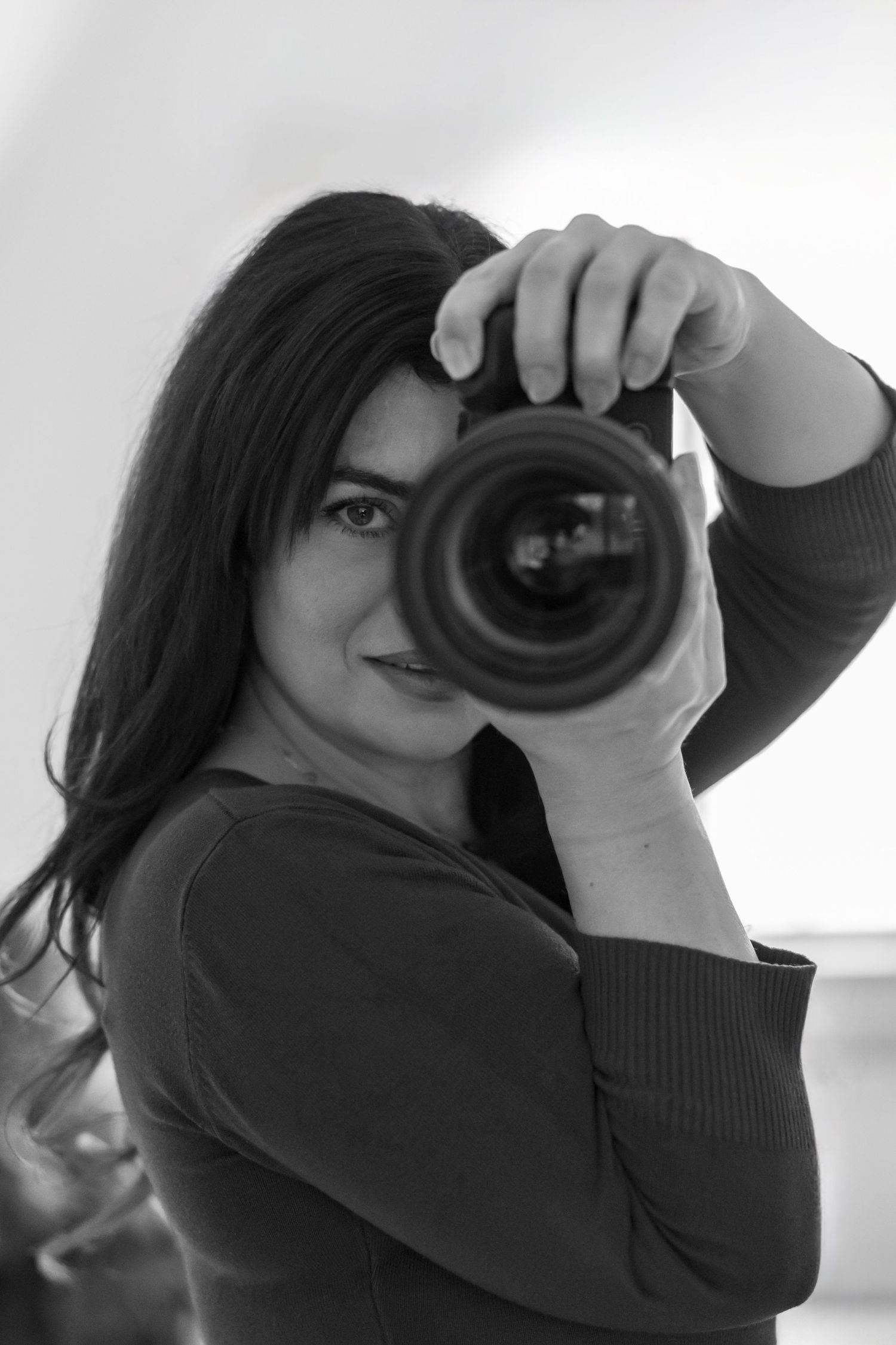 A photographer in a dark sweater holds up a professional camera with a large lens in black and white.