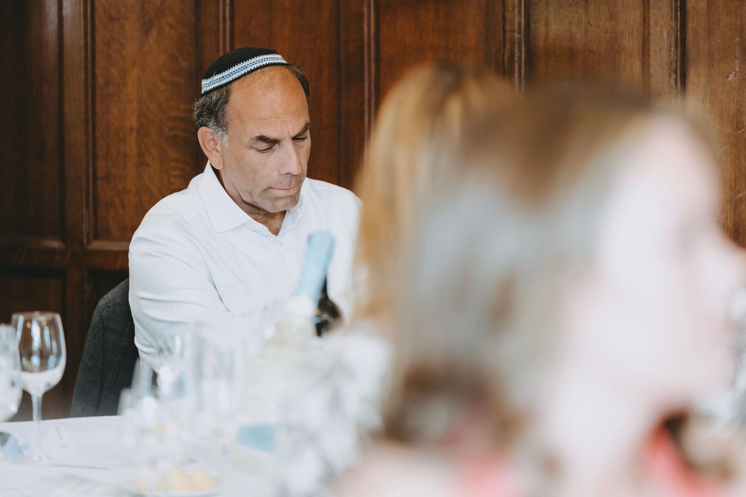 Wedding guest in yarmulke seated at reception table with wine glasses and place settings.