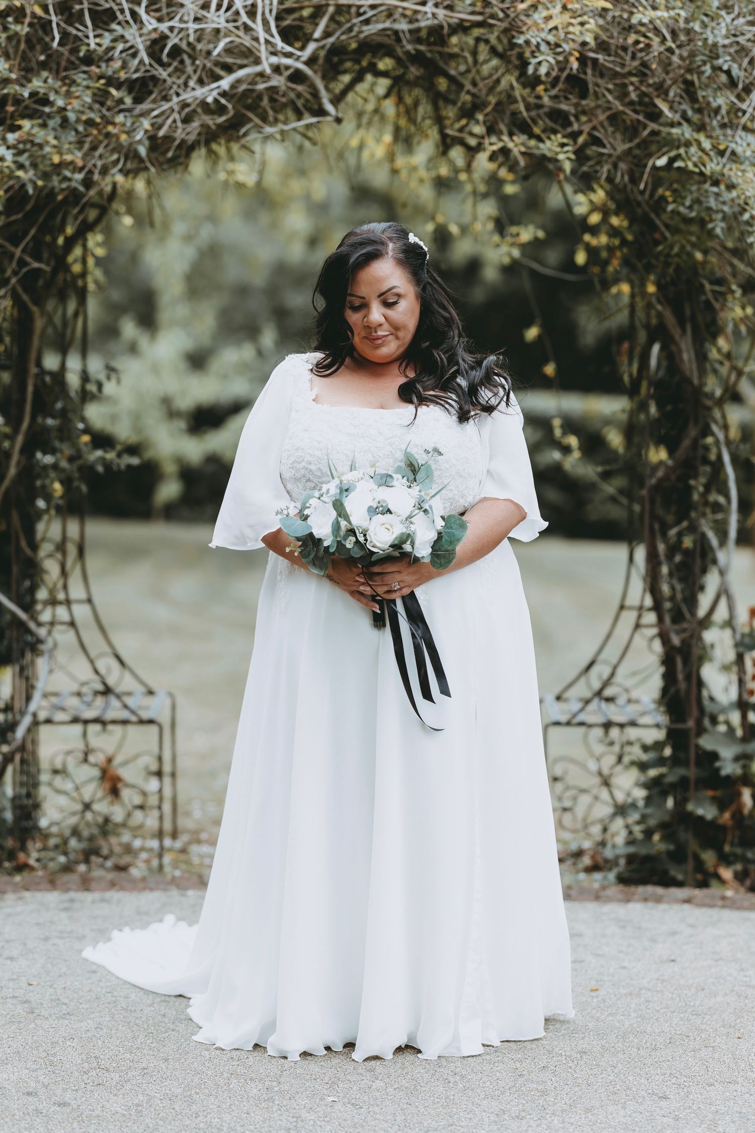 Bride in flowing white dress holds elegant white and green bouquet in garden setting.