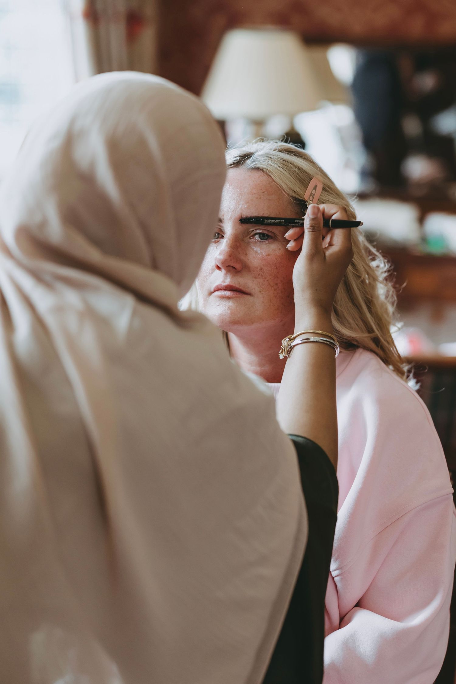 A makeup artist wearing a hijab applies eyebrow makeup to a blonde woman in a pink top.