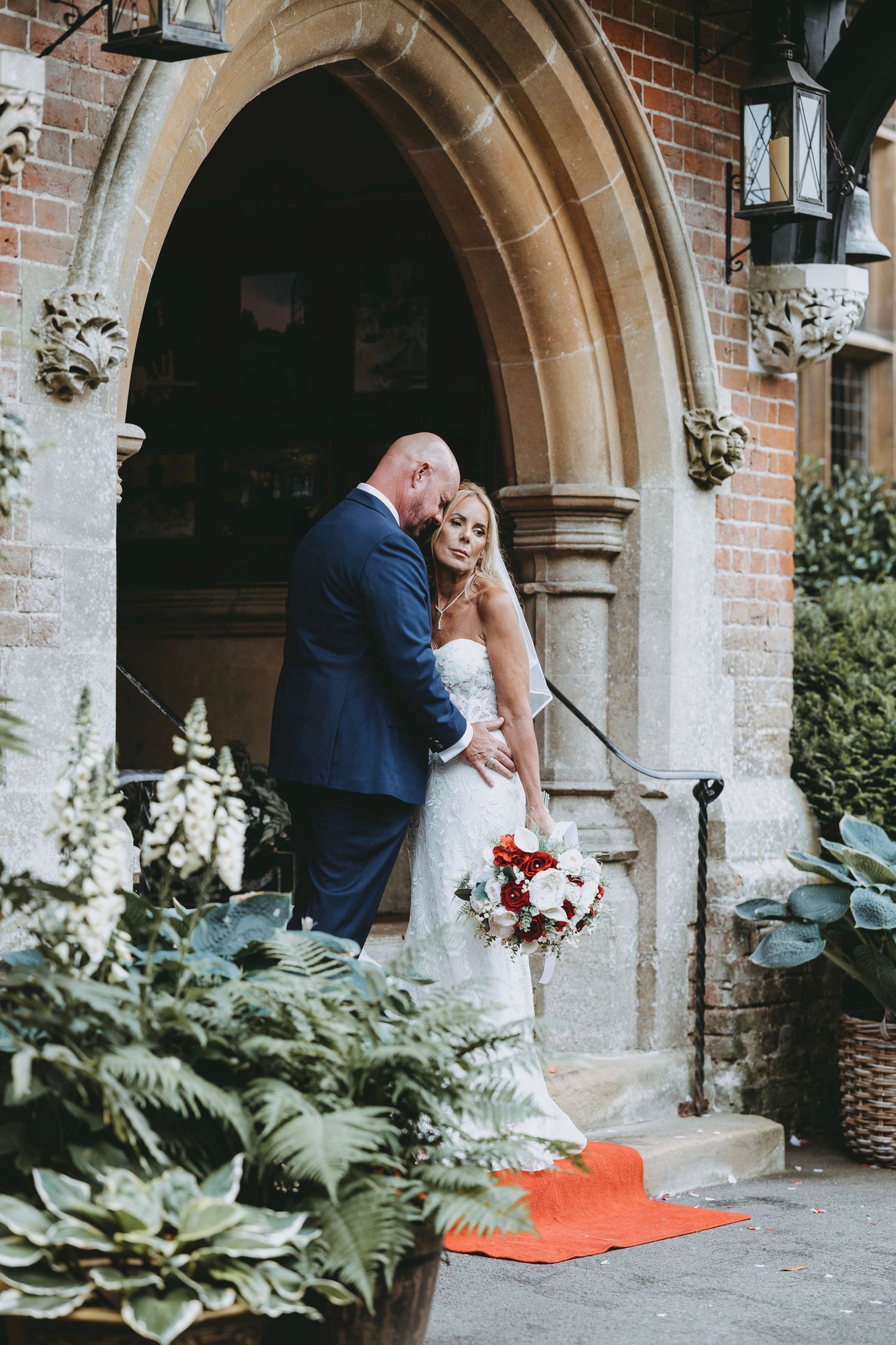 Bride and groom share a romantic moment outside a stone church entrance, bride holding a red and white bouquet.