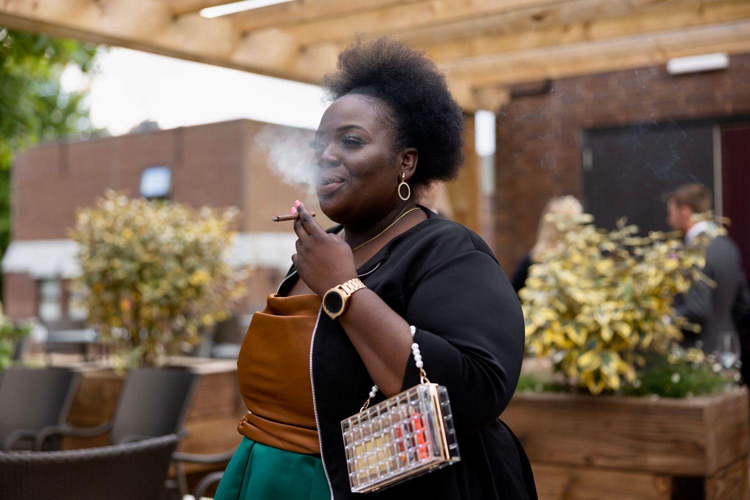 Person in a black top and green skirt smoking on an outdoor patio with decorative plants and wooden beams.