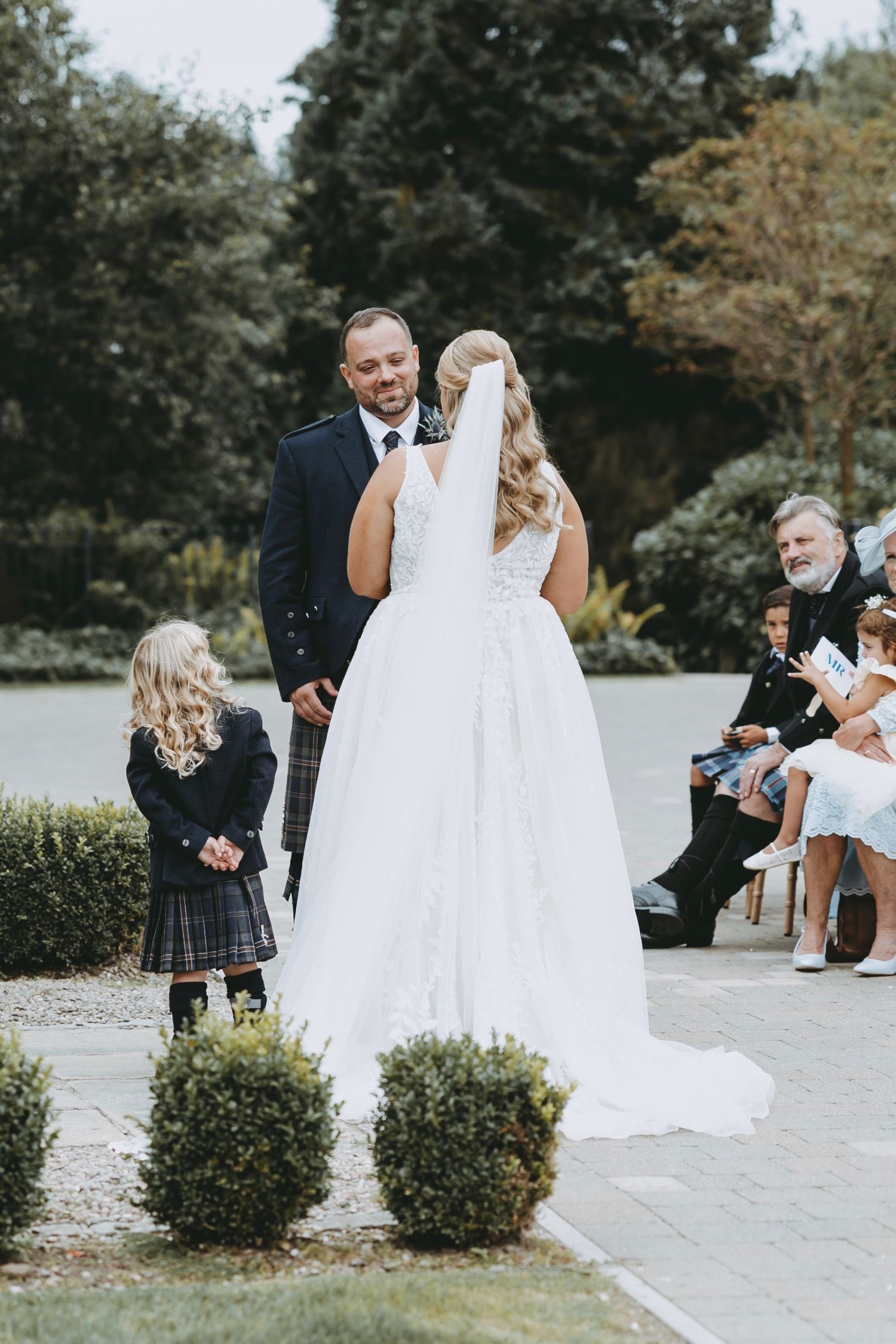 Wedding ceremony scene with decorative shrubs and guests seated outdoors.