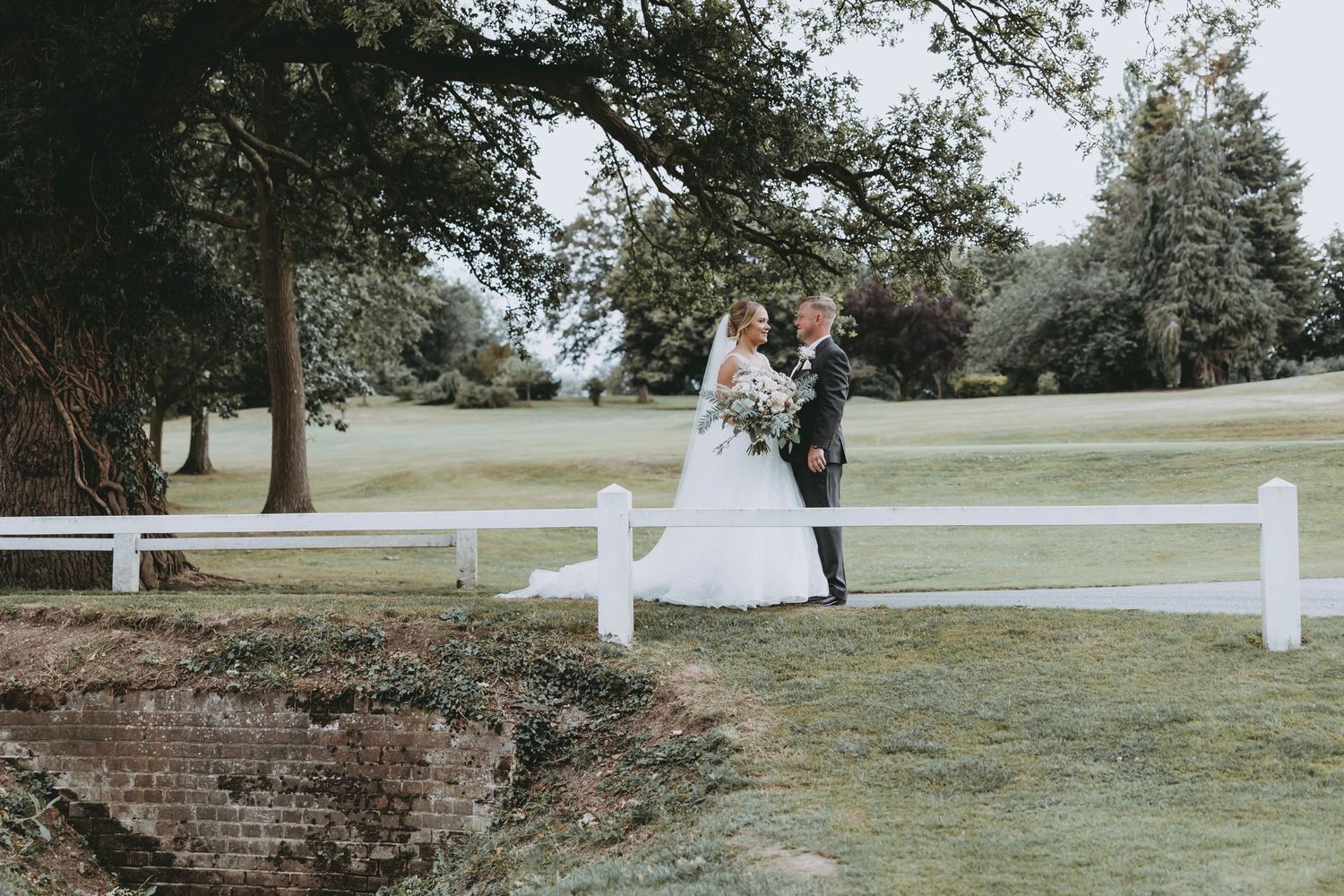Newlyweds posing by white fence on scenic golf course.