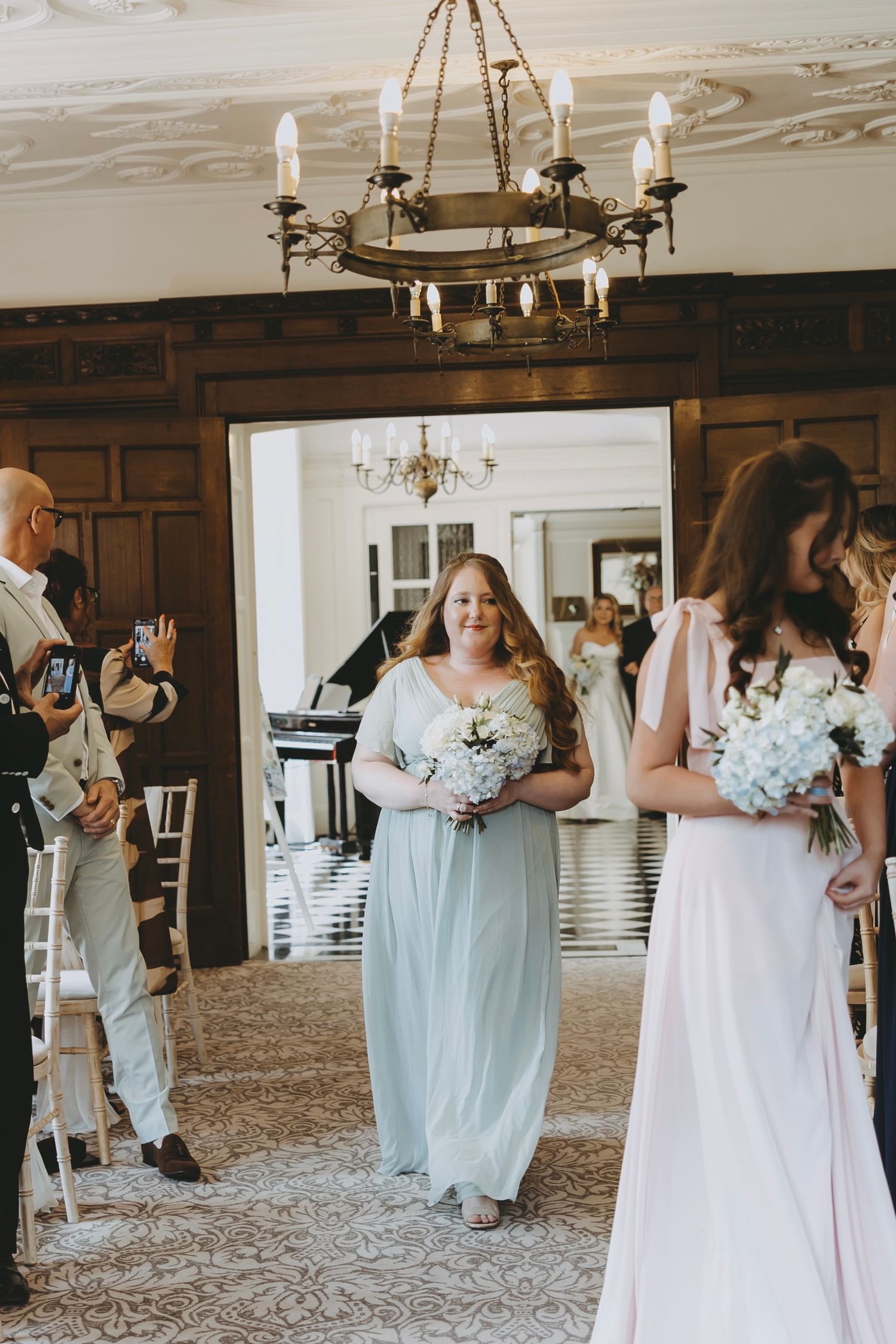 Wedding ceremony in progress inside historic venue with dark wood walls and ornate chandelier.