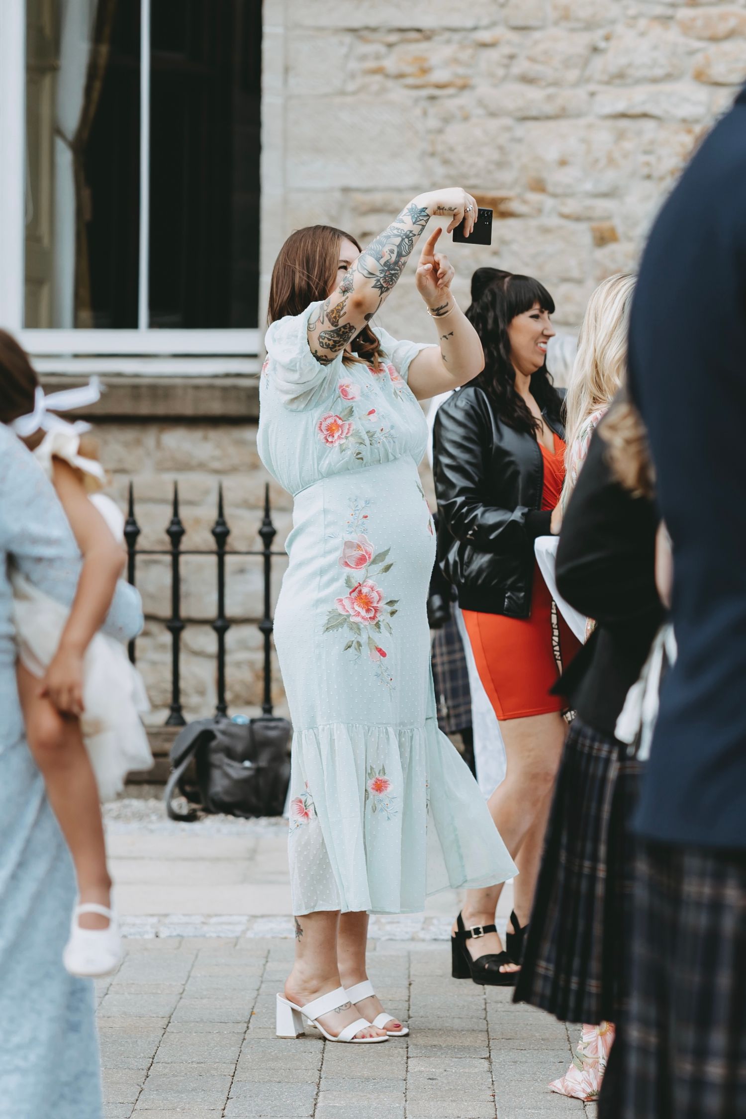 Wedding guests in formal attire mingling outside stone building.