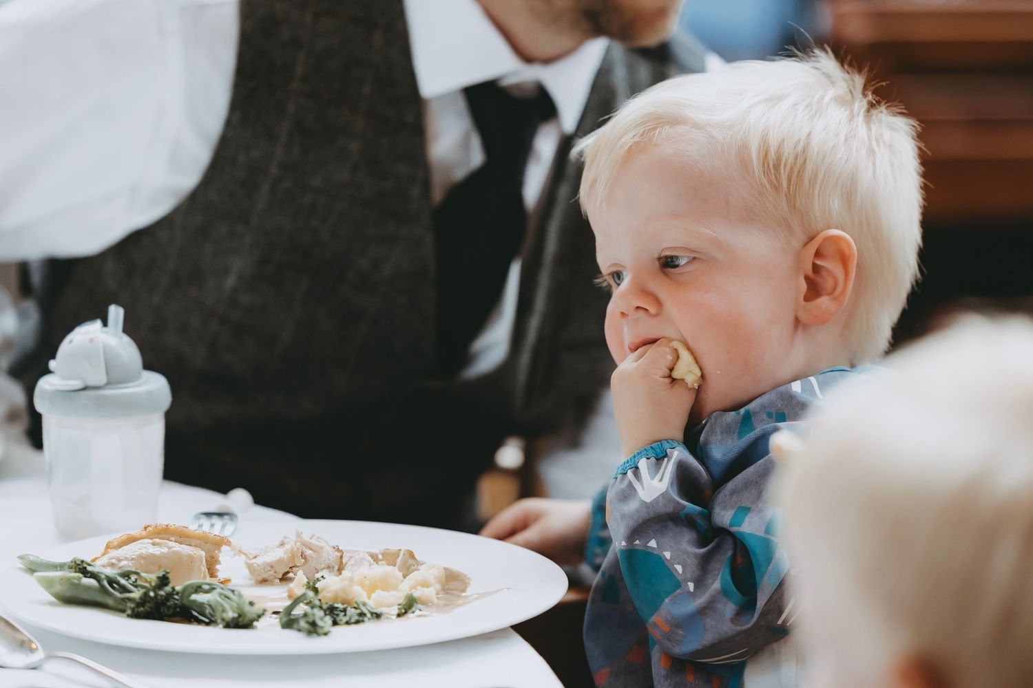 A young child in formal wear smiles while seated at a dining table with a plate of food.
