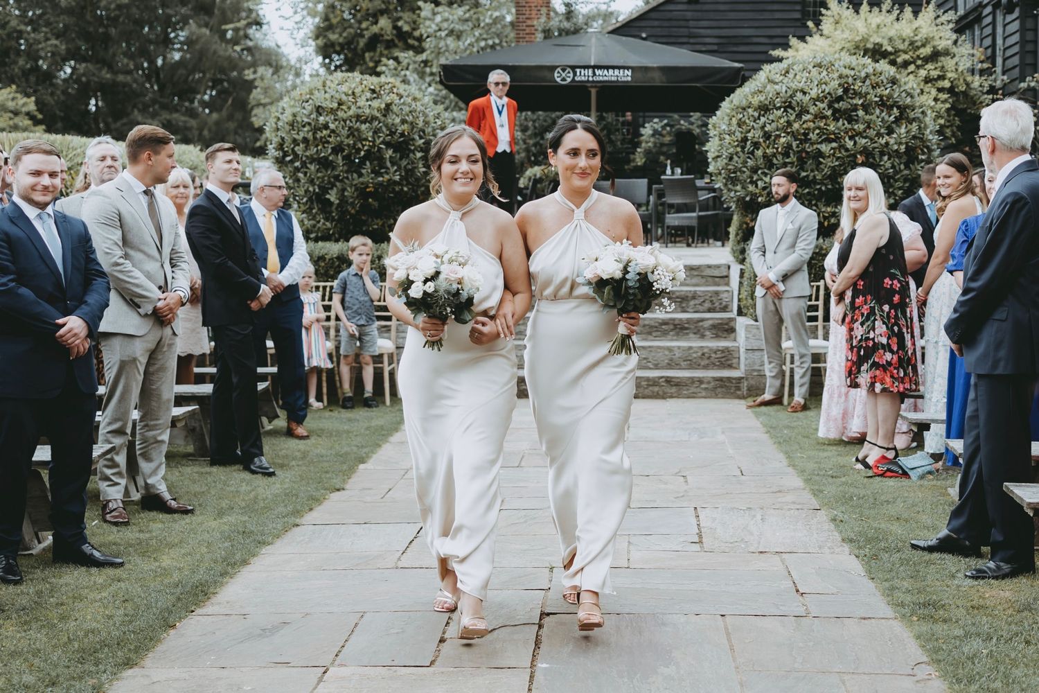 Wedding couple walks down stone aisle carrying bouquets while guests watch from both sides.