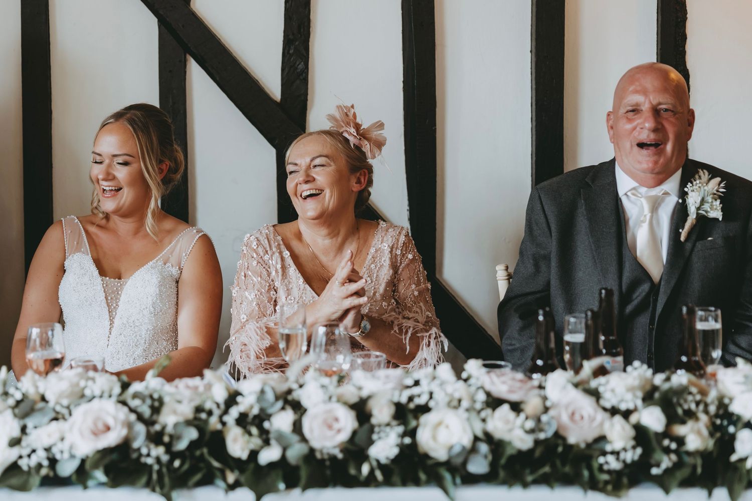 Wedding party members laugh and smile while seated at a head table decorated with white floral arrangements.